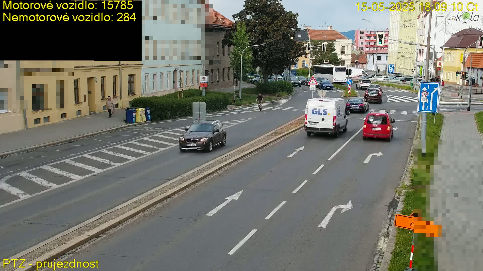 Several cars and a van are driving on a multi-lane road in front of buildings on a partly sunny day.