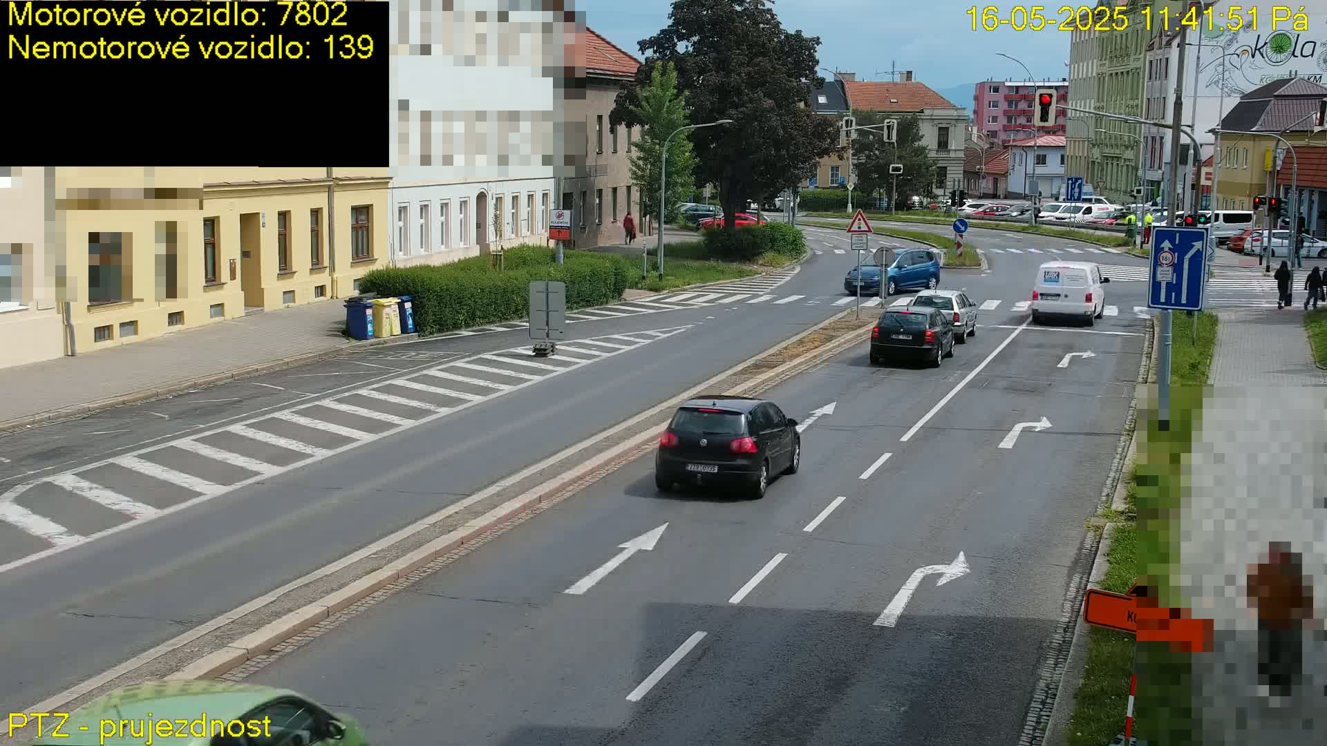 Several cars are driving on a paved road in front of buildings on a partly sunny day.