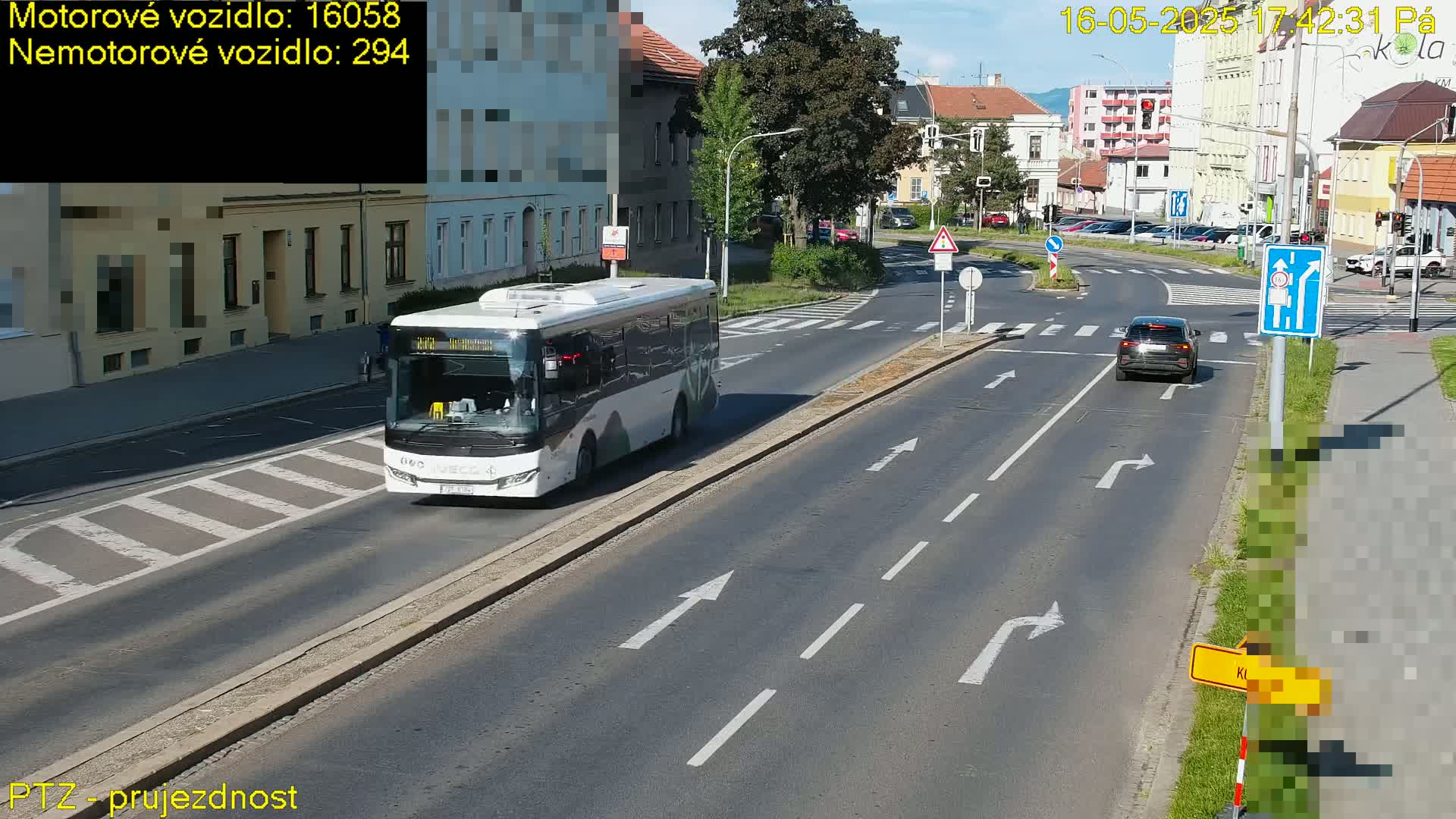 A city street scene on a sunny day shows a bus and a car driving on a multi-lane road with pedestrian crossings,  surrounded by buildings.