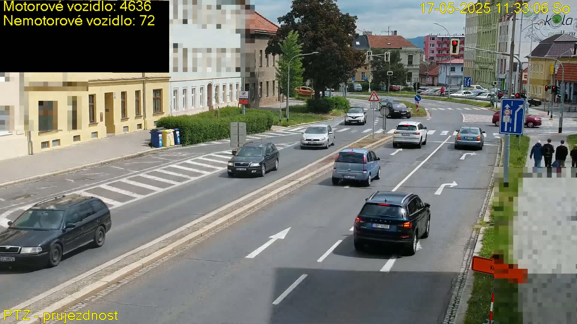 Several cars are driving on a multi-lane road on a sunny day, with pedestrians walking on the sidewalk.