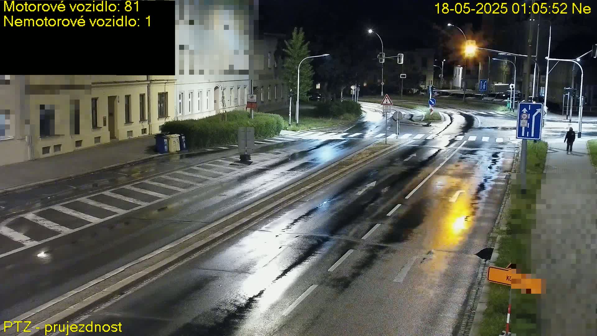 A wet, empty road intersection at night, lit by streetlights, with a few buildings visible in the background.