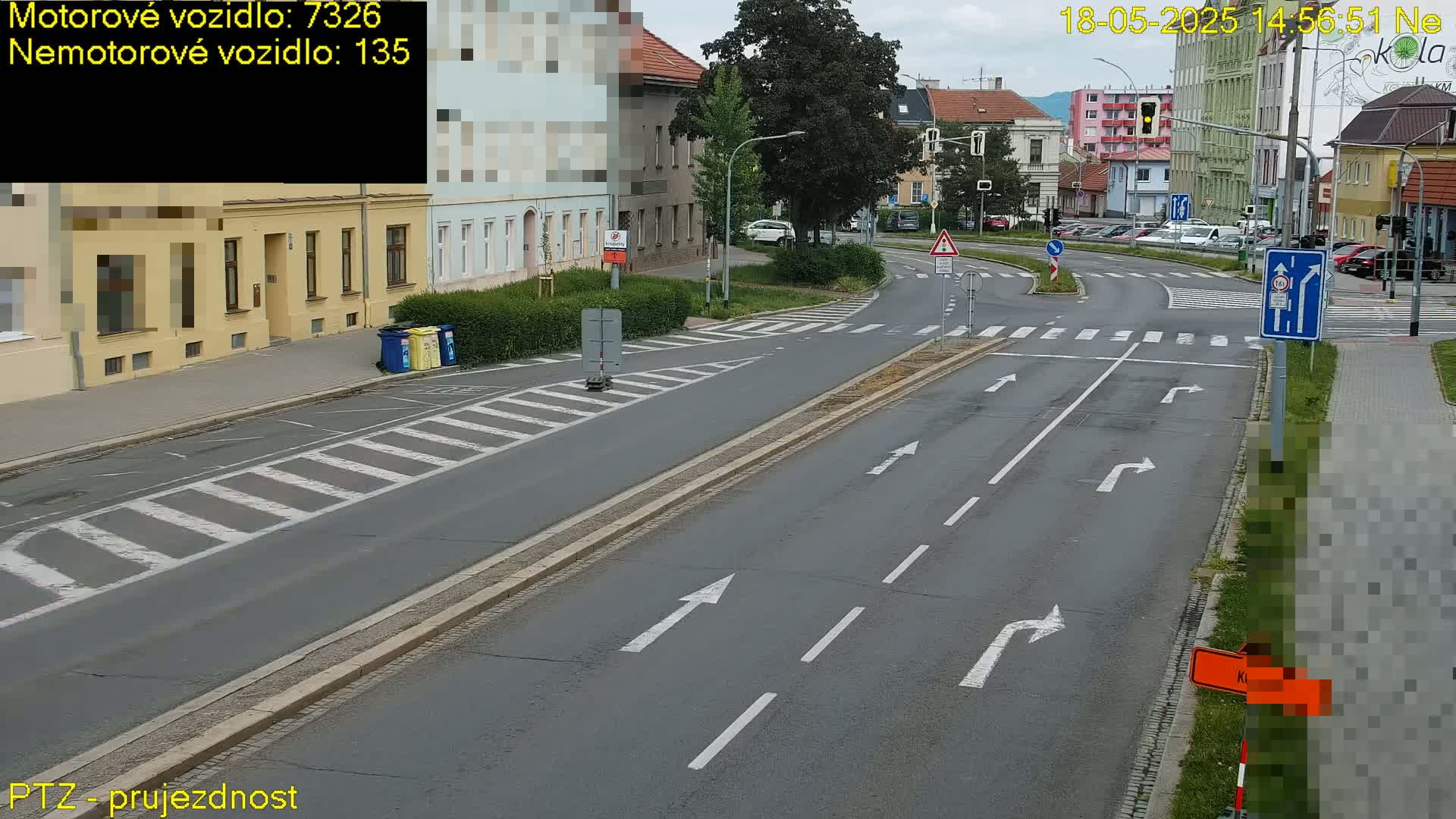 A mostly empty road intersection with several buildings and cars visible in the background on a partly cloudy day.