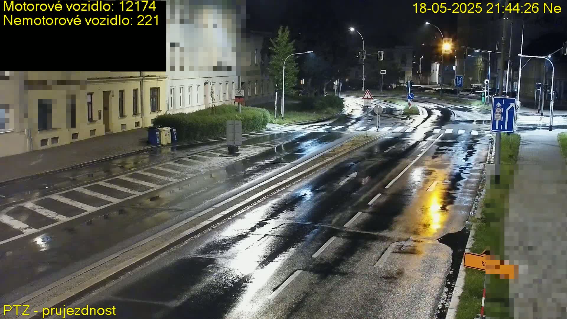 A wet, nighttime street scene shows a road intersection with streetlights, buildings, and pedestrian crossings under a dark sky.