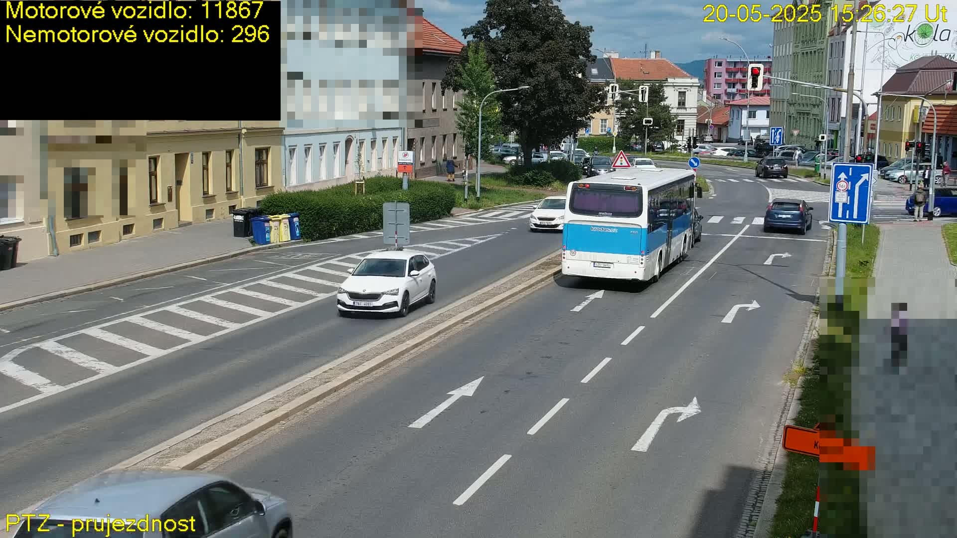 A sunny day on a city street shows a white car, a blue bus, and other vehicles at an intersection with pedestrian crosswalks.