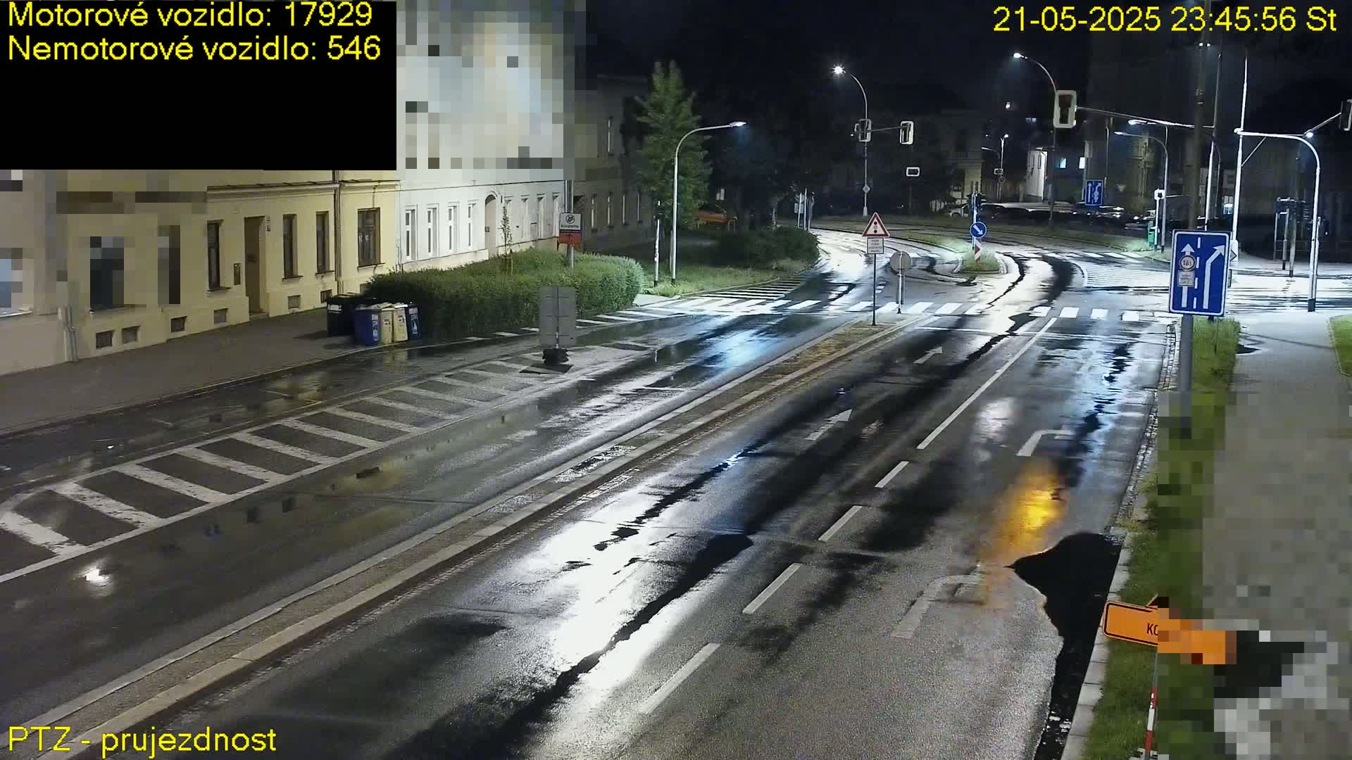 A wet, empty road at night, illuminated by streetlights, with buildings lining one side and some patchy vegetation on the other.