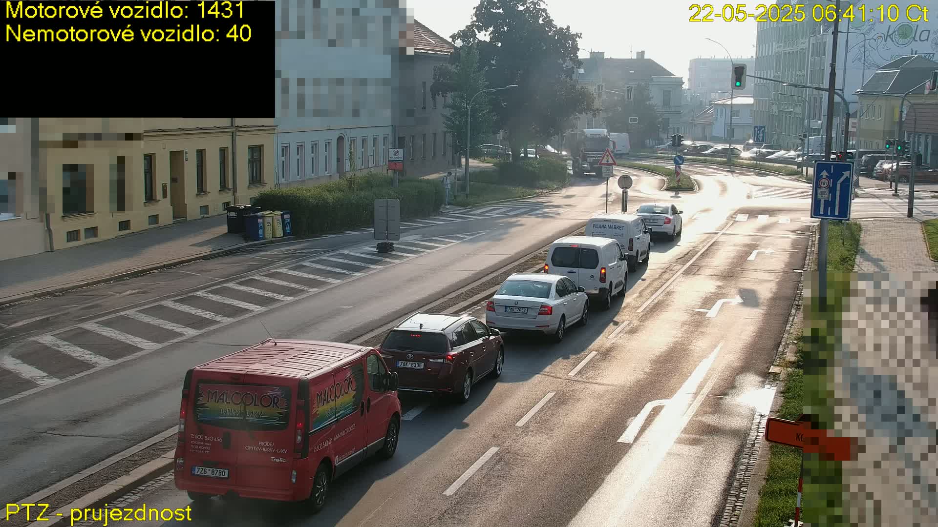 A sunny day shows a line of cars stopped at an intersection in front of buildings; a red van is at the very front of the line.