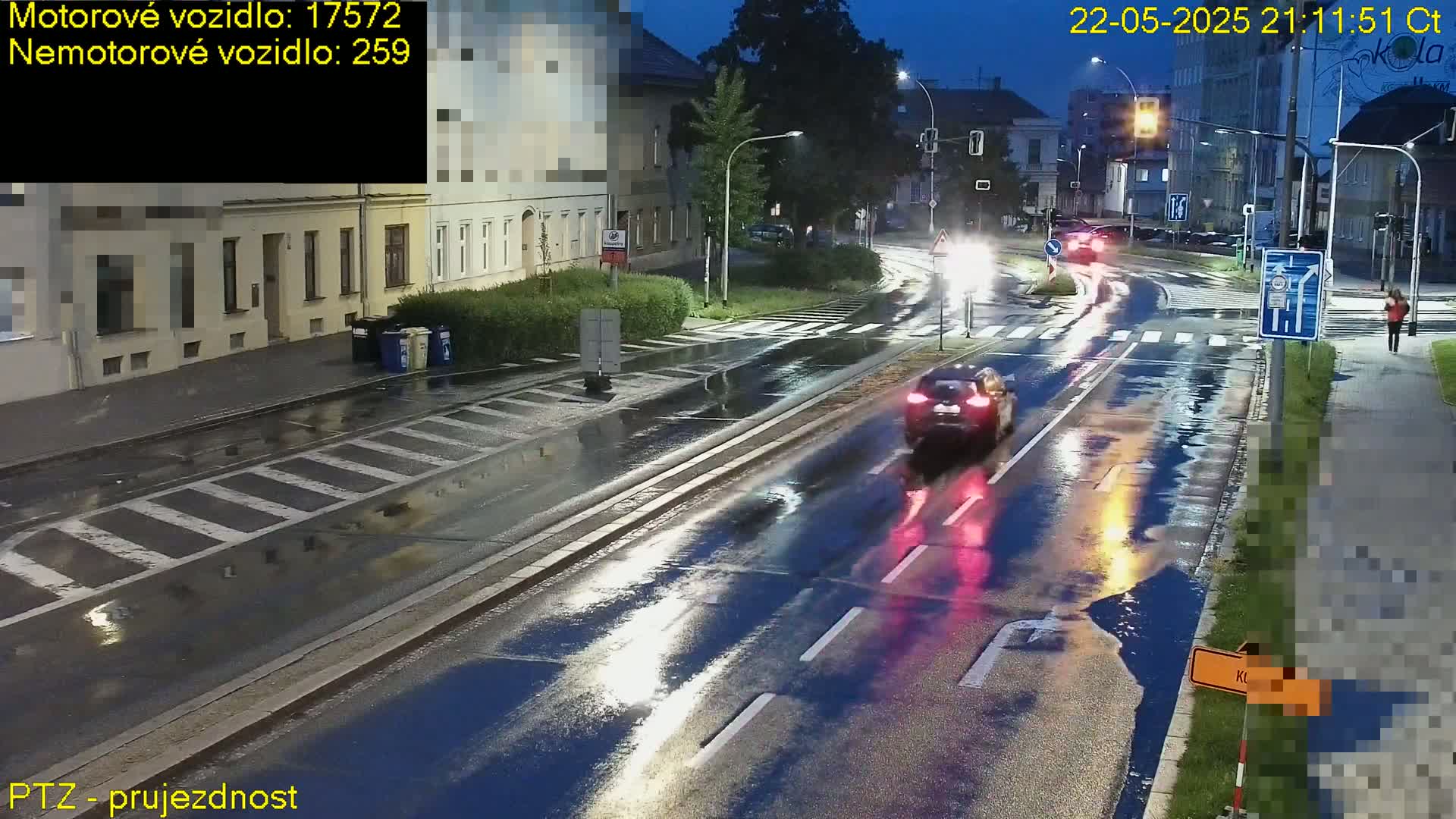 A wet, nighttime street scene shows cars driving through a crossroads in front of buildings.