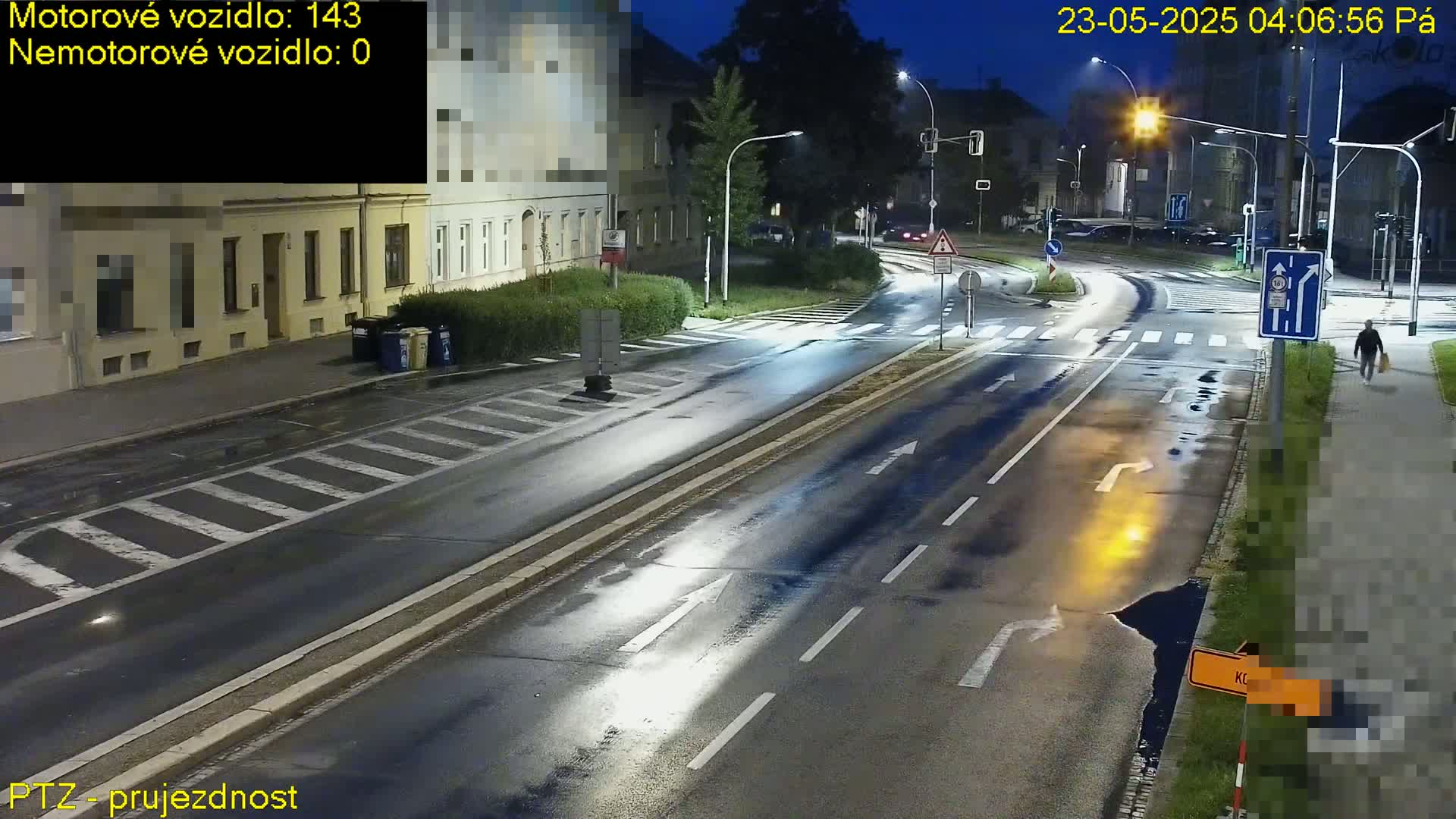 A wet, empty road intersection at night, lit by streetlights, with a pedestrian walking on the sidewalk.