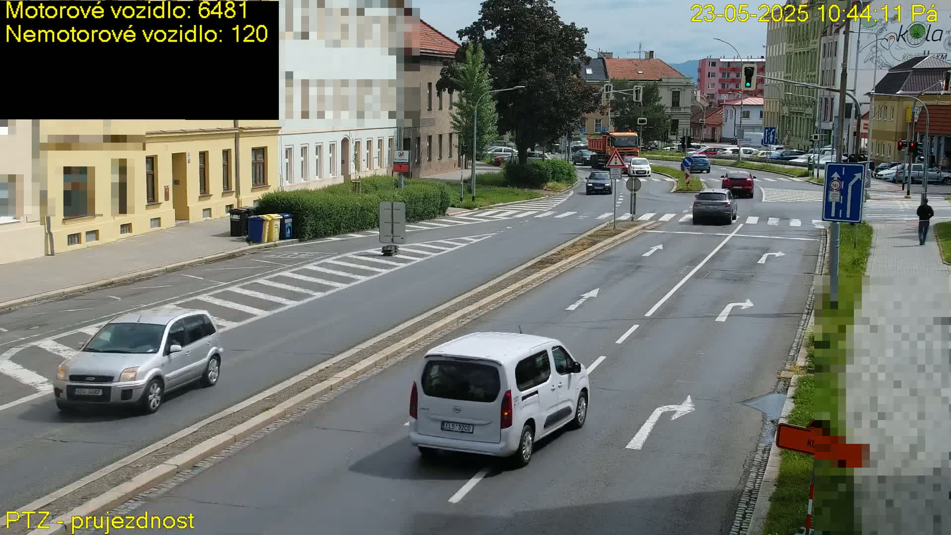 A sunny day shows several cars driving on a multi-lane road that intersects with a circular road, surrounded by buildings and pedestrians.