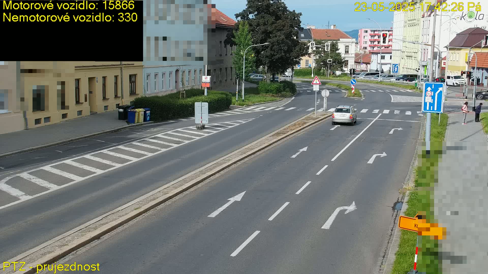 A sunny day shows a multi-lane road with a car driving through an intersection near buildings, pedestrian crosswalks, and some people on the sidewalk.
