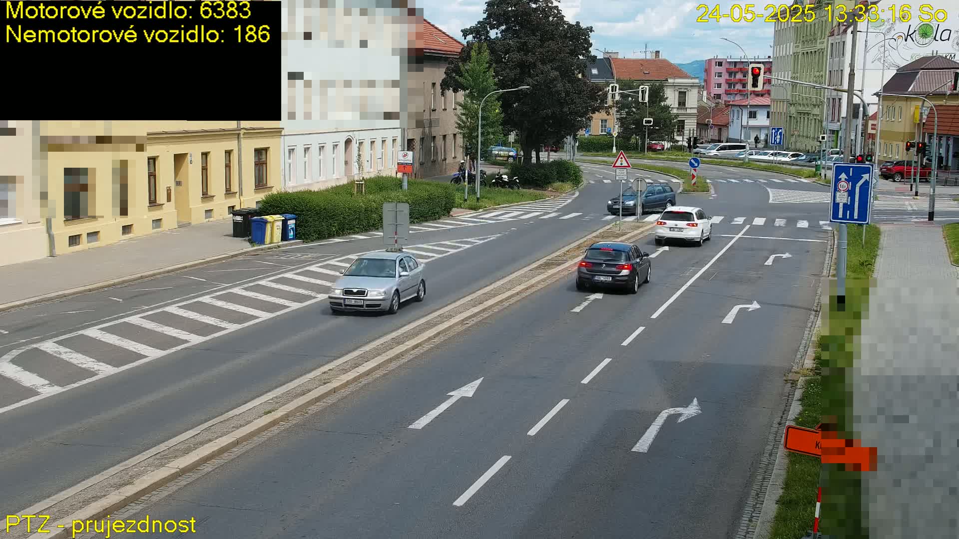 Several cars are driving on a multi-lane road on a sunny day, with buildings and trees visible in the background.