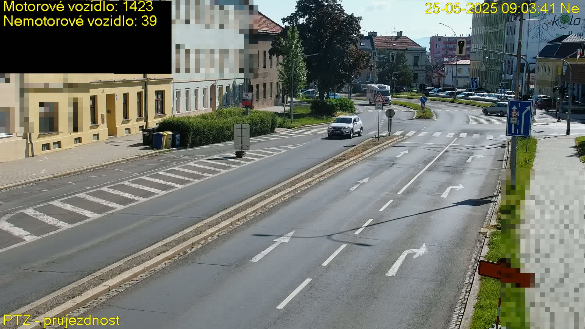 A sunny day shows a multi-lane road with a white SUV driving and several other vehicles at an intersection, with buildings and greenery lining the road.