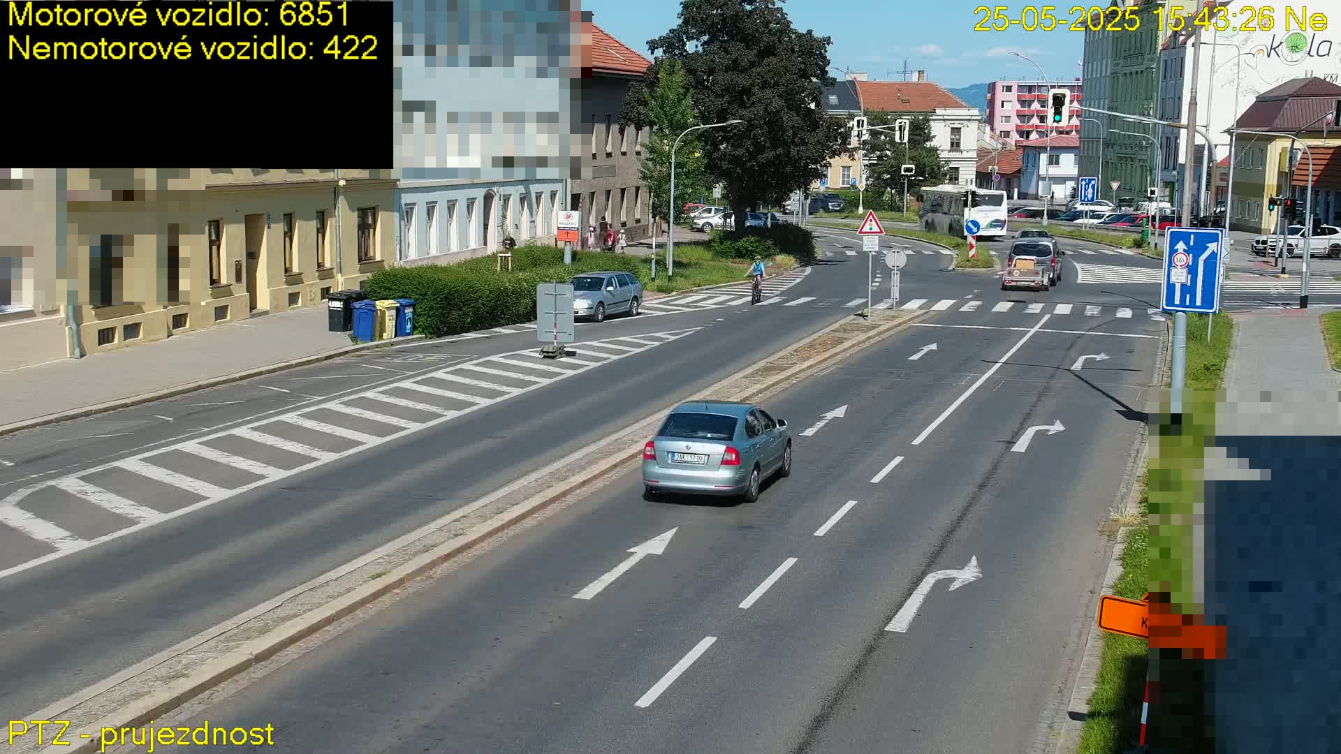 A sunny day reveals a multi-lane road with several cars and a cyclist at an intersection, with buildings lining the street.