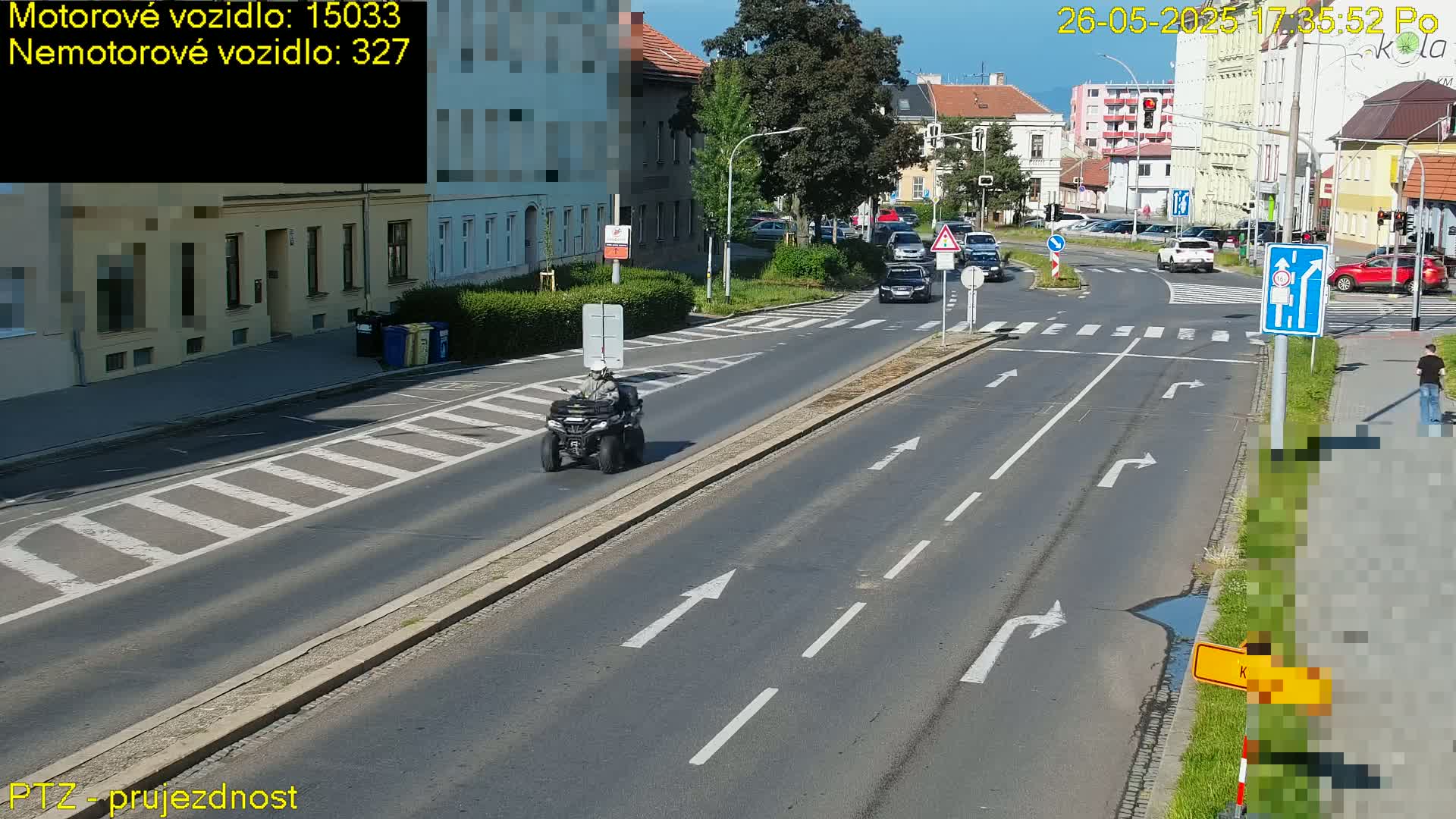 A four-wheeler ATV travels down a multi-lane road on a sunny day, with cars at an intersection in the background.