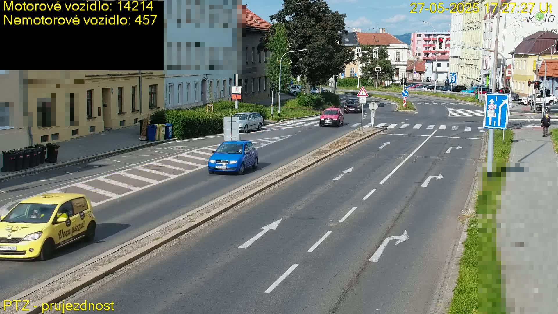 A sunny day shows a multi-lane road with several cars and a pedestrian, situated in a town with buildings lining the street.