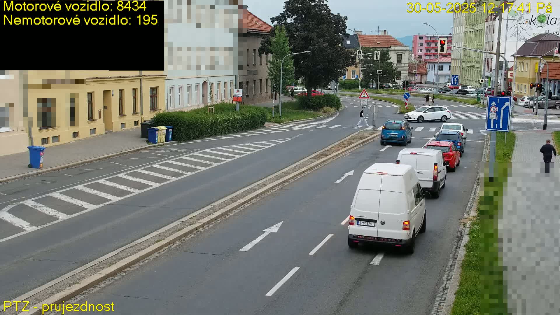A street scene on a sunny day shows several vehicles, including vans and cars, driving on a road with pedestrian crossings and buildings lining the street.