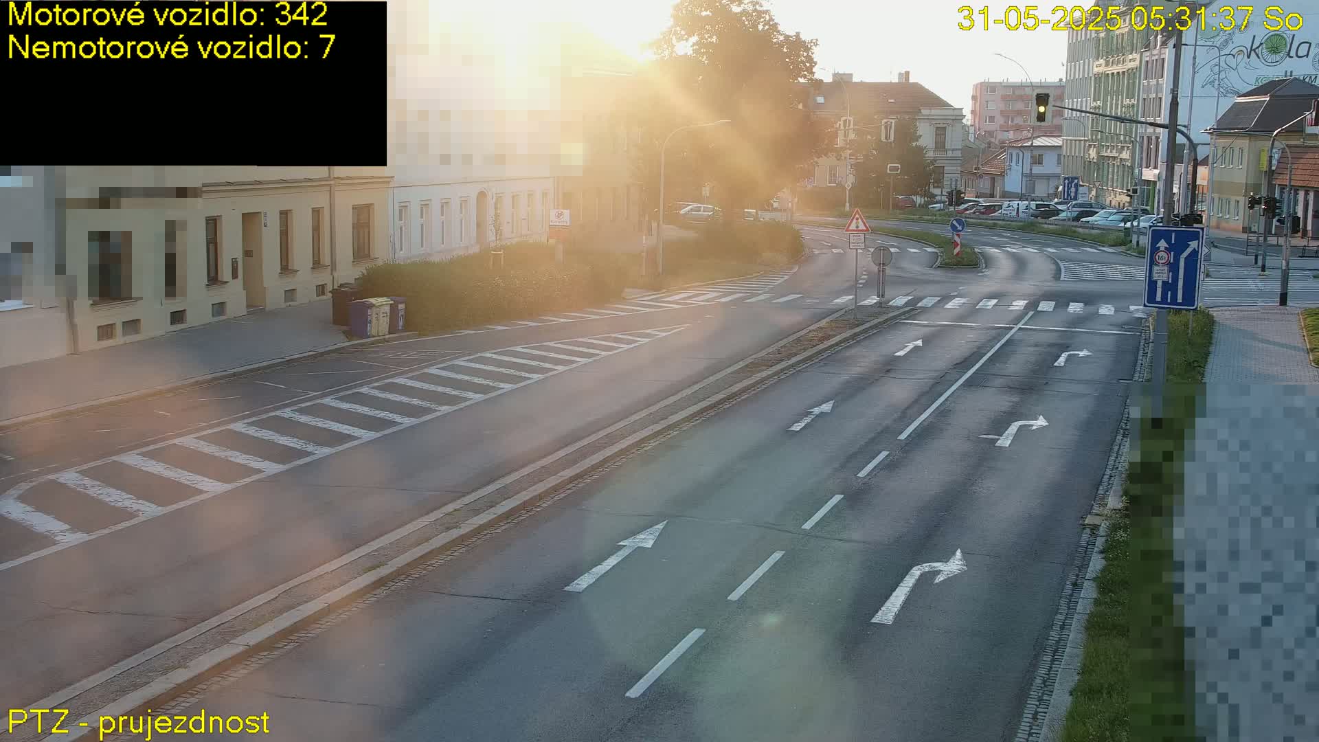 A sunny morning view of a multi-lane road intersection with buildings and parked cars visible in the background.