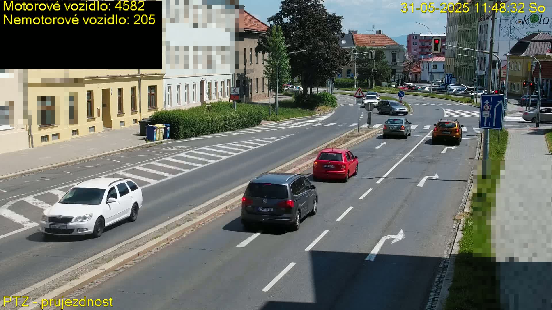 Several cars are driving on a multi-lane road on a sunny day, with buildings and pedestrian crossings visible.
