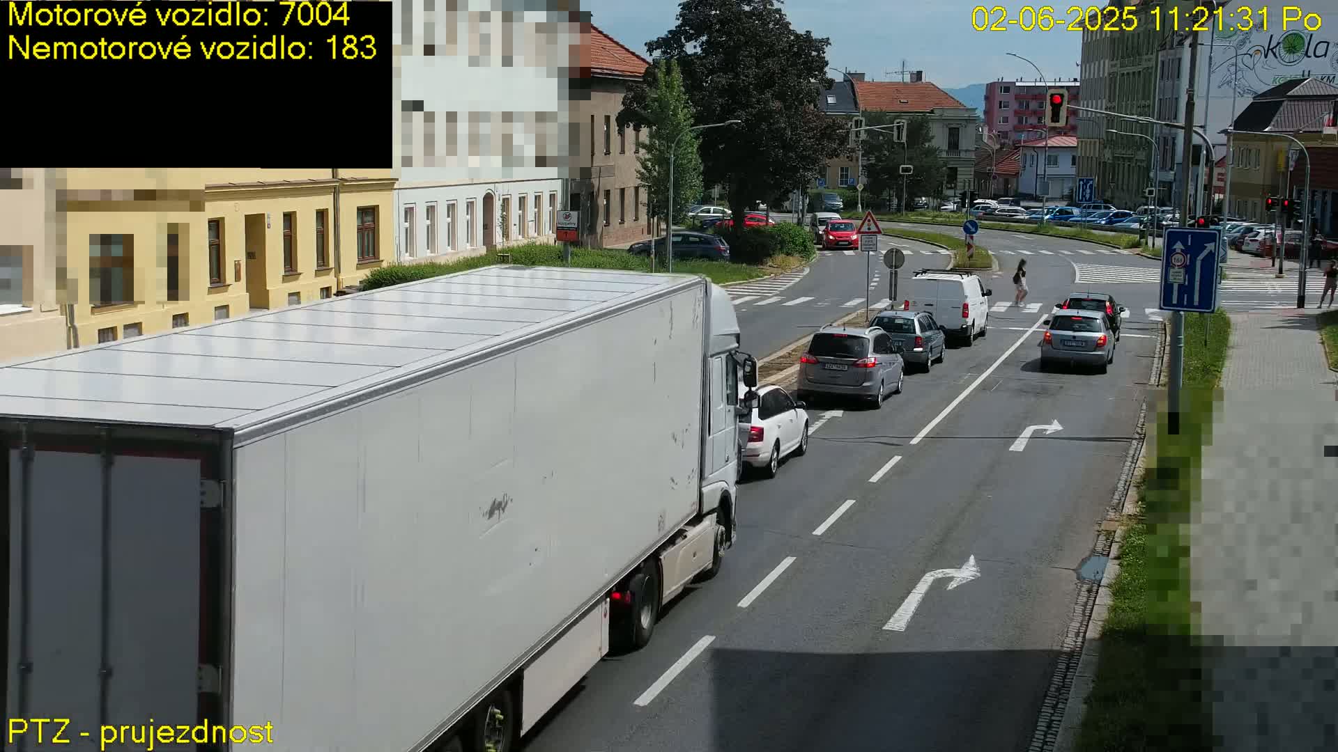 A large white semi-truck is driving on a road next to several cars and a pedestrian crossing a street on a sunny day.
