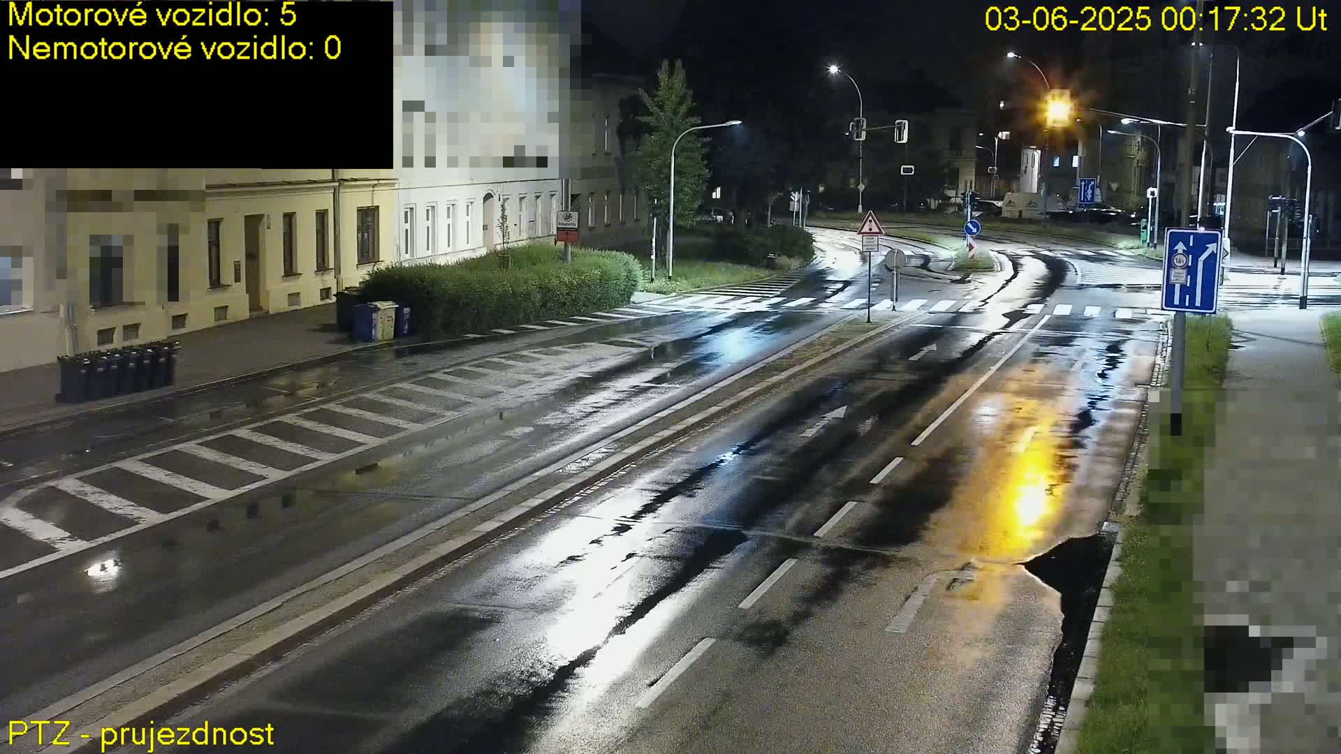 A wet, empty street at night, illuminated by streetlights, with buildings lining one side.