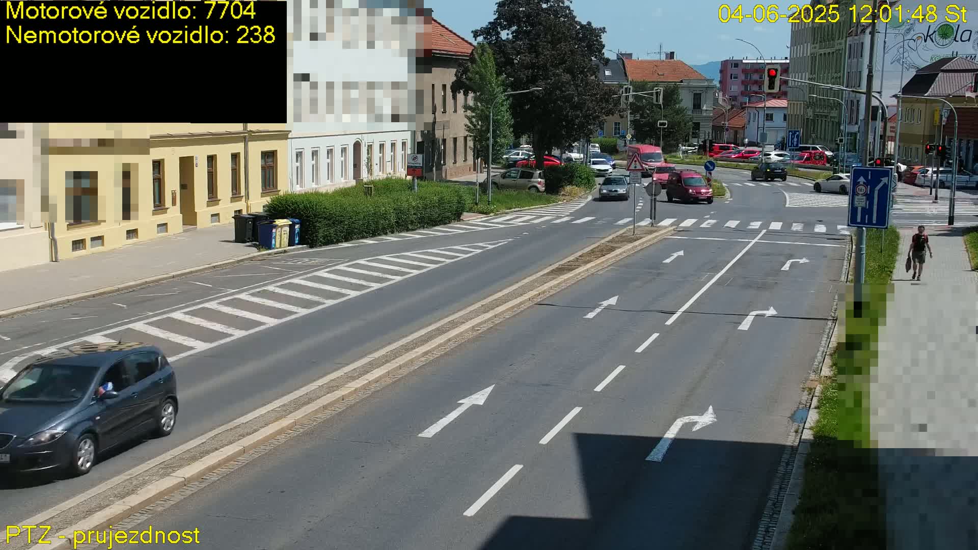 A sunny day shows a multi-lane road intersection with several cars and a pedestrian walking on a sidewalk.