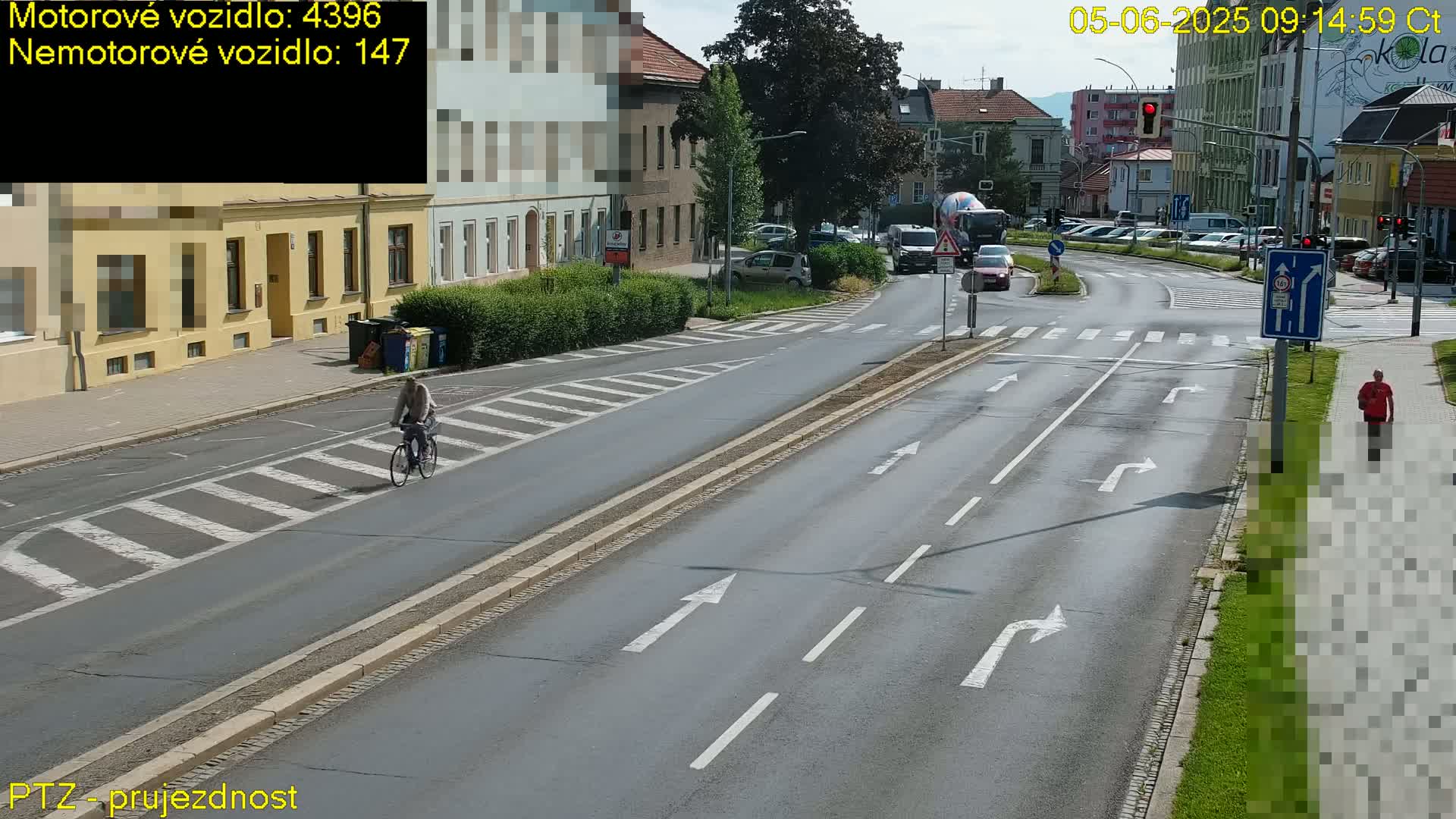 A cyclist rides along a multi-lane road on a sunny day, with cars and pedestrians visible at a nearby intersection.