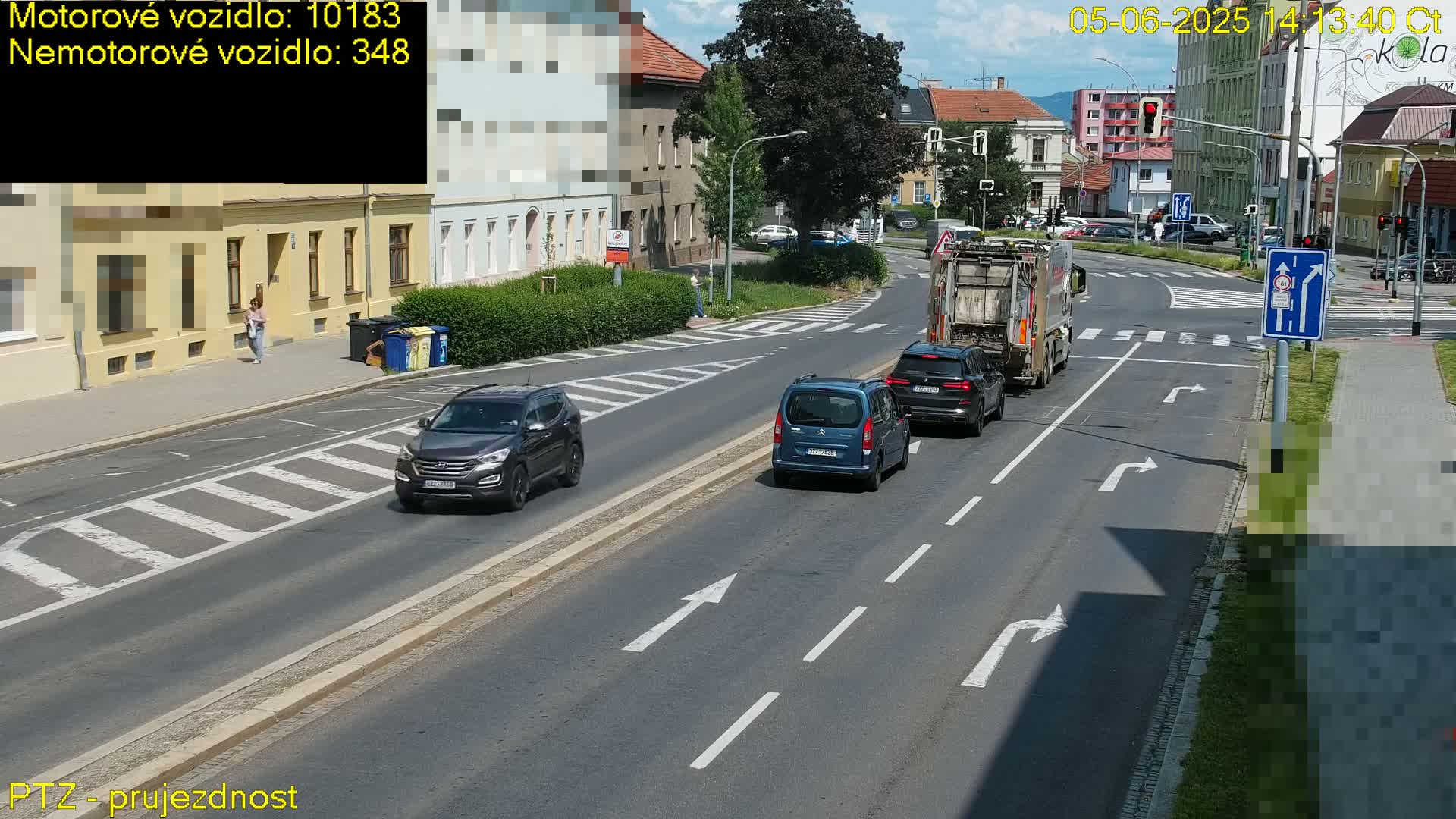 On a sunny day, several cars, a van, and a garbage truck are driving on a multi-lane road that intersects with another road at a crosswalk where a pedestrian is walking.