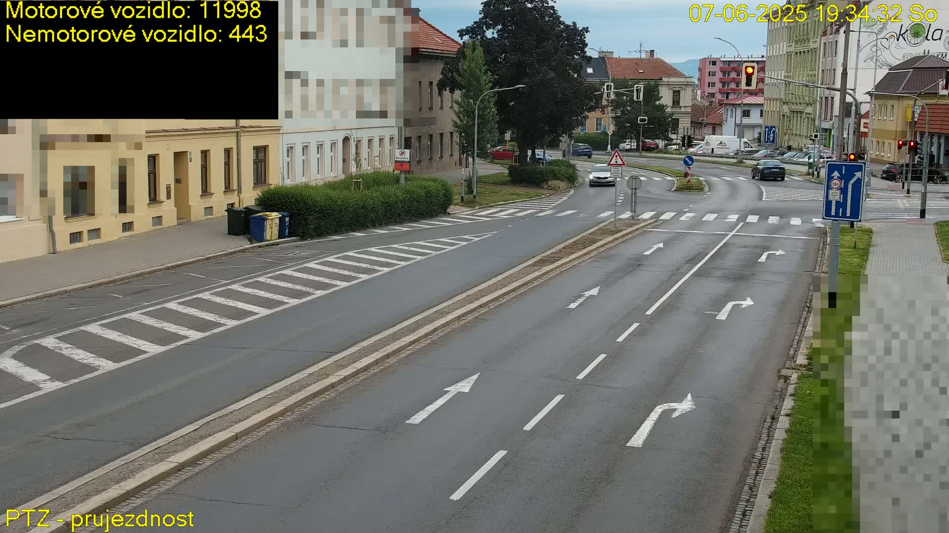 A wet, multi-lane road with white lane markings and a crosswalk intersects with a roundabout, surrounded by buildings and greenery under an overcast sky.