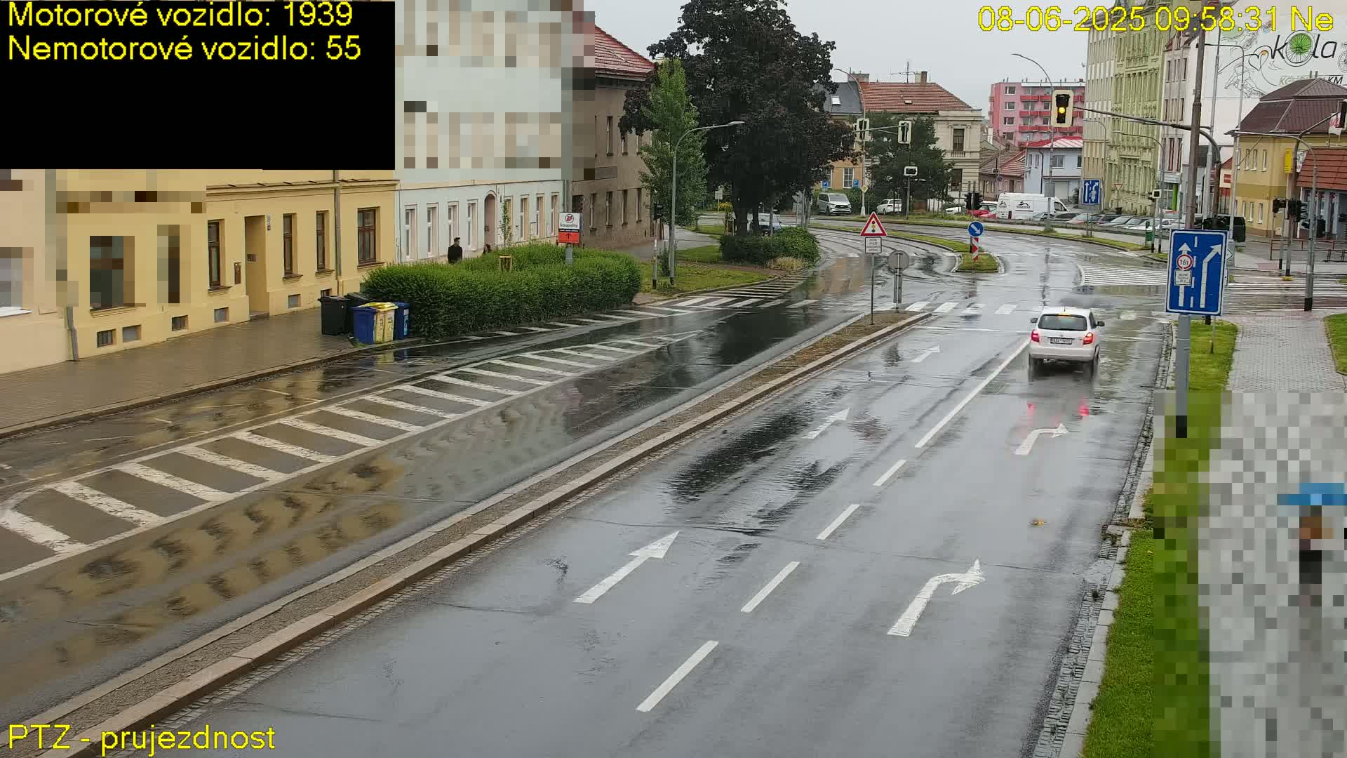 A wet, rainy street scene shows a small white car driving through an intersection with buildings lining the road and pedestrian crosswalks.