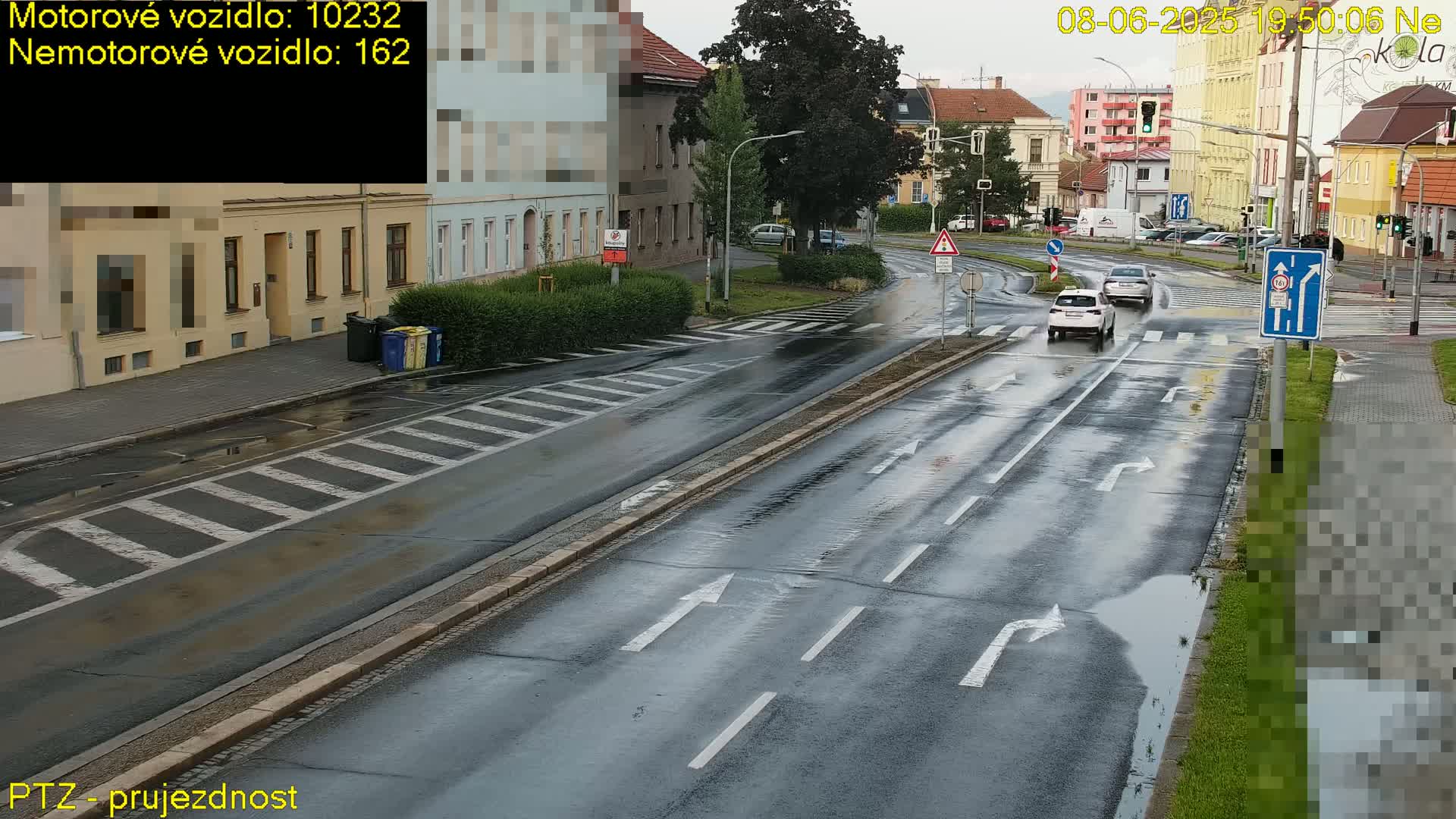 A wet, rain-slicked road with two cars driving through a city intersection, lined with buildings and greenery.