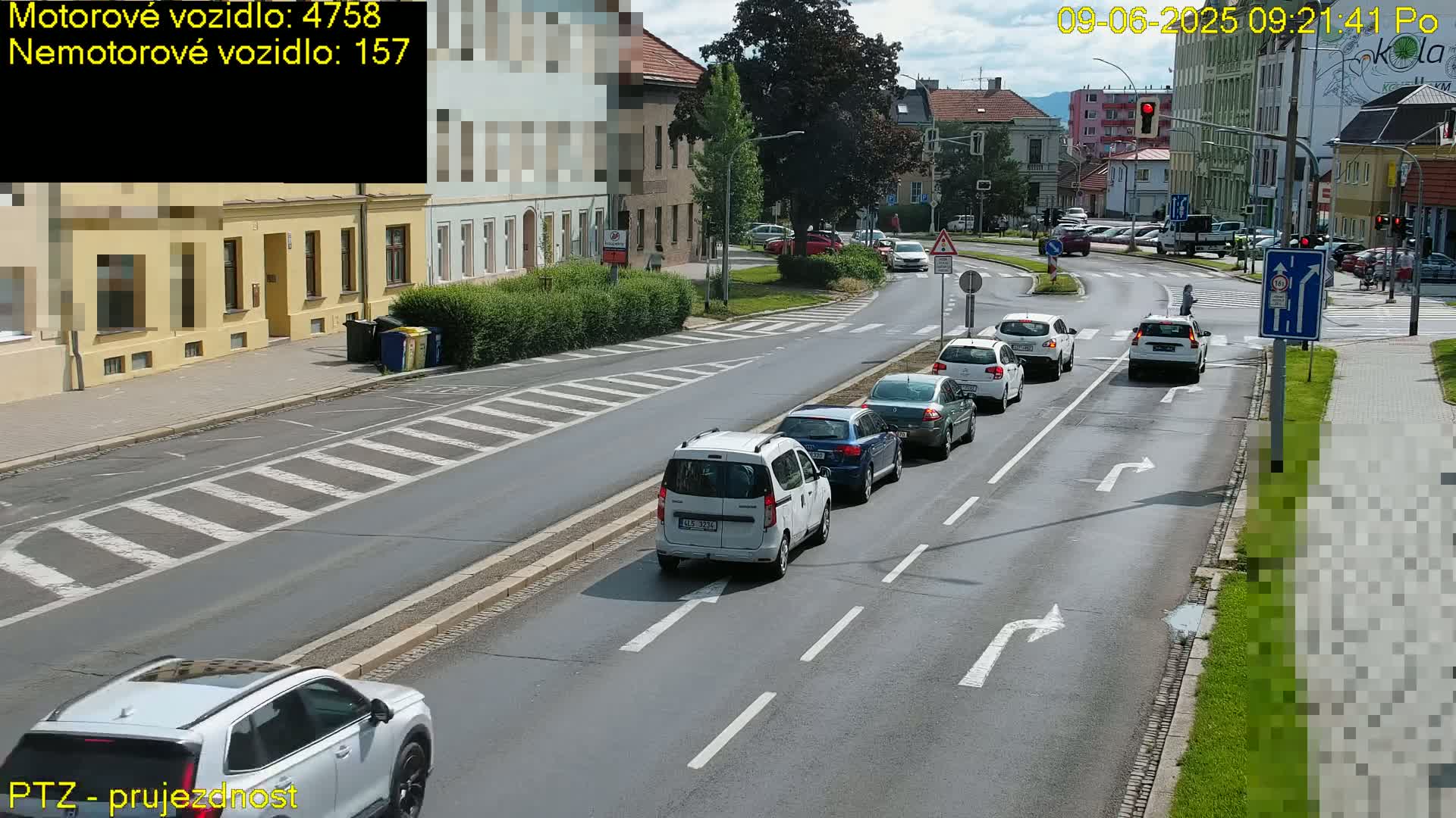 A sunny day view of a multi-lane road with several cars driving in both directions, passing through a pedestrian crossing in a town with buildings and greenery visible on the sides.