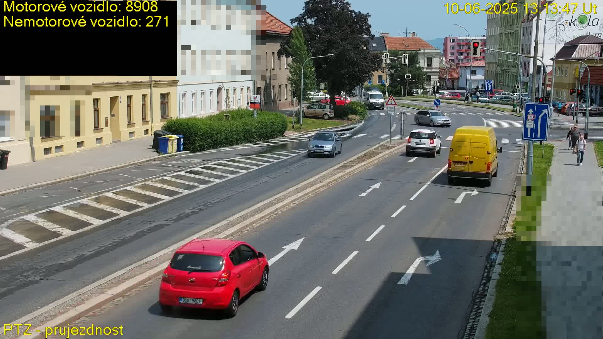 Several cars and a van are driving on a sunny city street, with pedestrians walking on a nearby sidewalk.