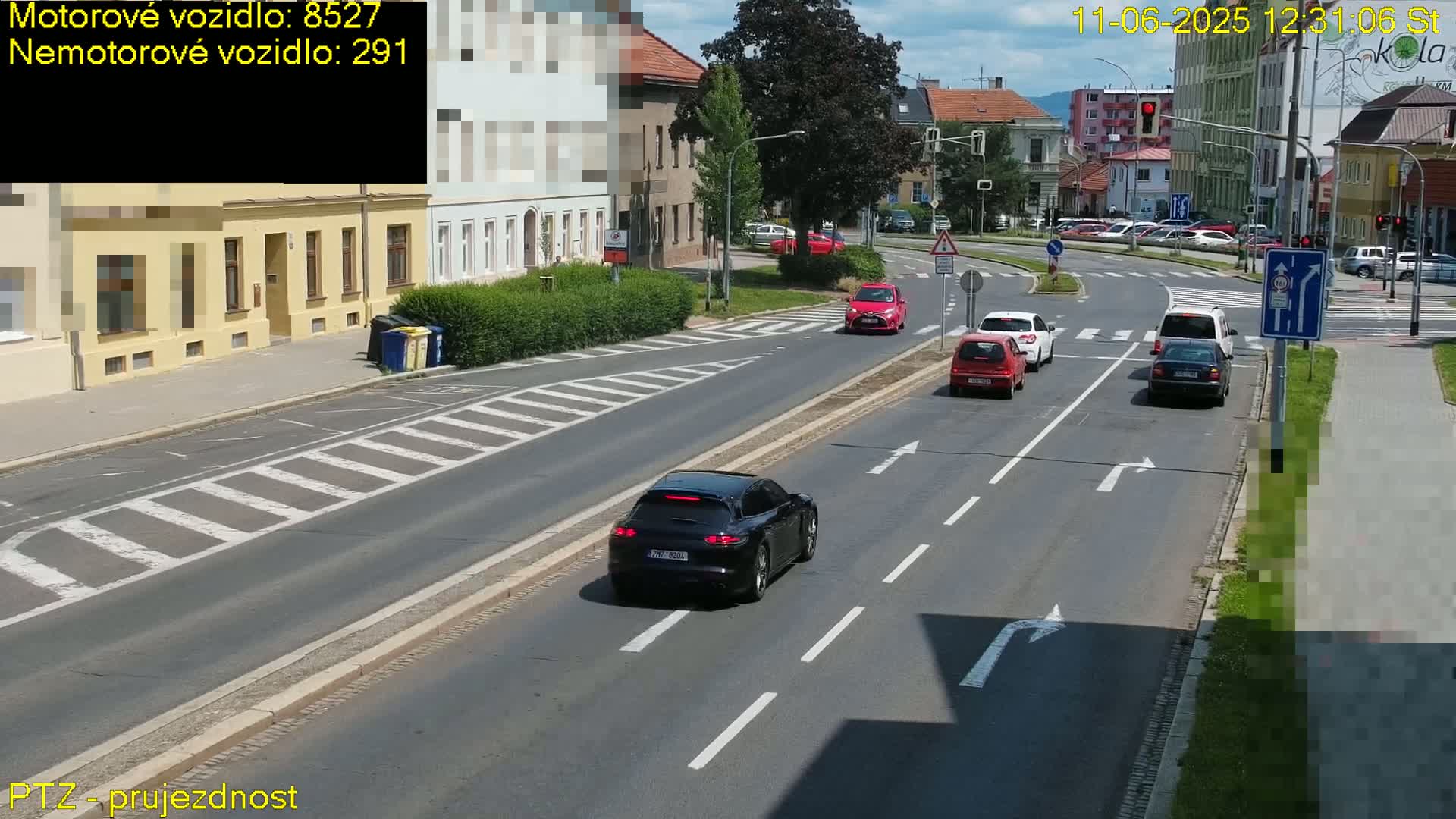 A black Porsche and several other cars are driving on a sunny street with buildings and pedestrian crossings visible.