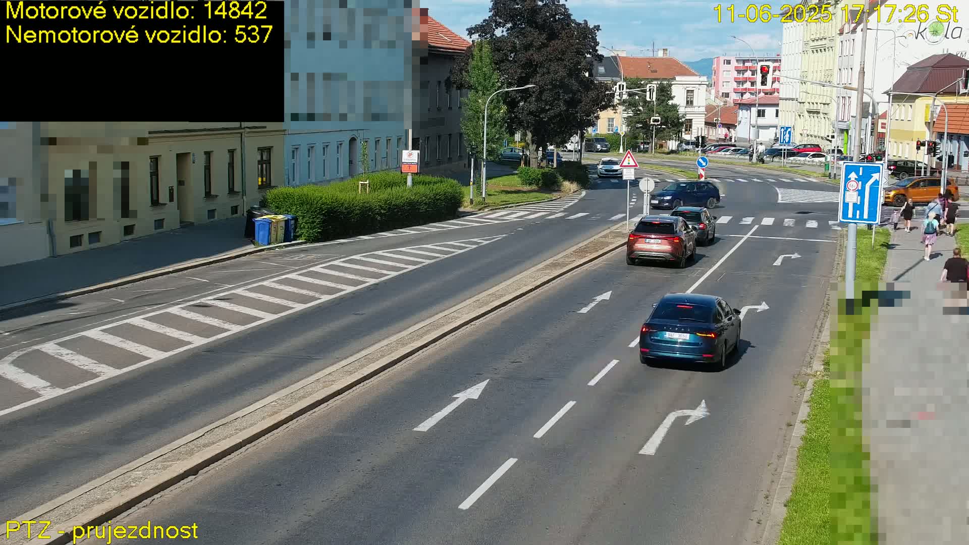 A sunny day on a multi-lane road with several cars driving, pedestrians walking on a sidewalk, and buildings lining the street.