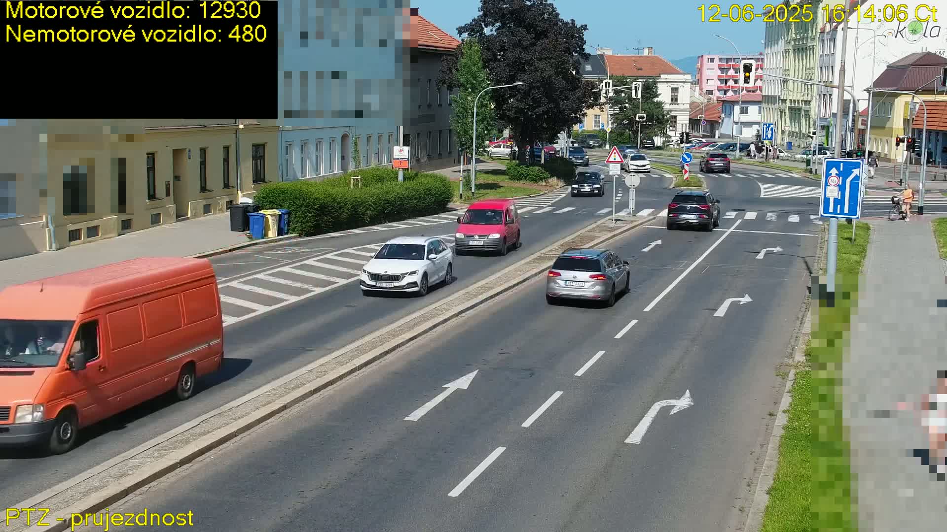 Several cars and a van are driving on a sunny road that intersects with another road, with a pedestrian walking on a sidewalk next to the road and buildings lining the street.