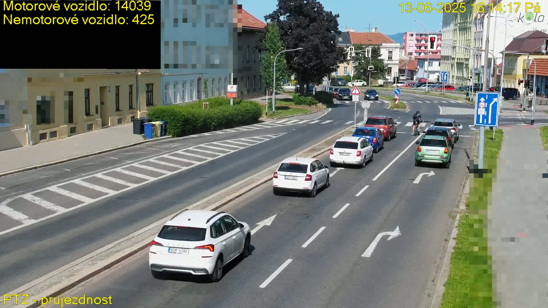Several white cars and a light green SUV are driving on a sunny street lined with buildings, with a cyclist riding alongside.