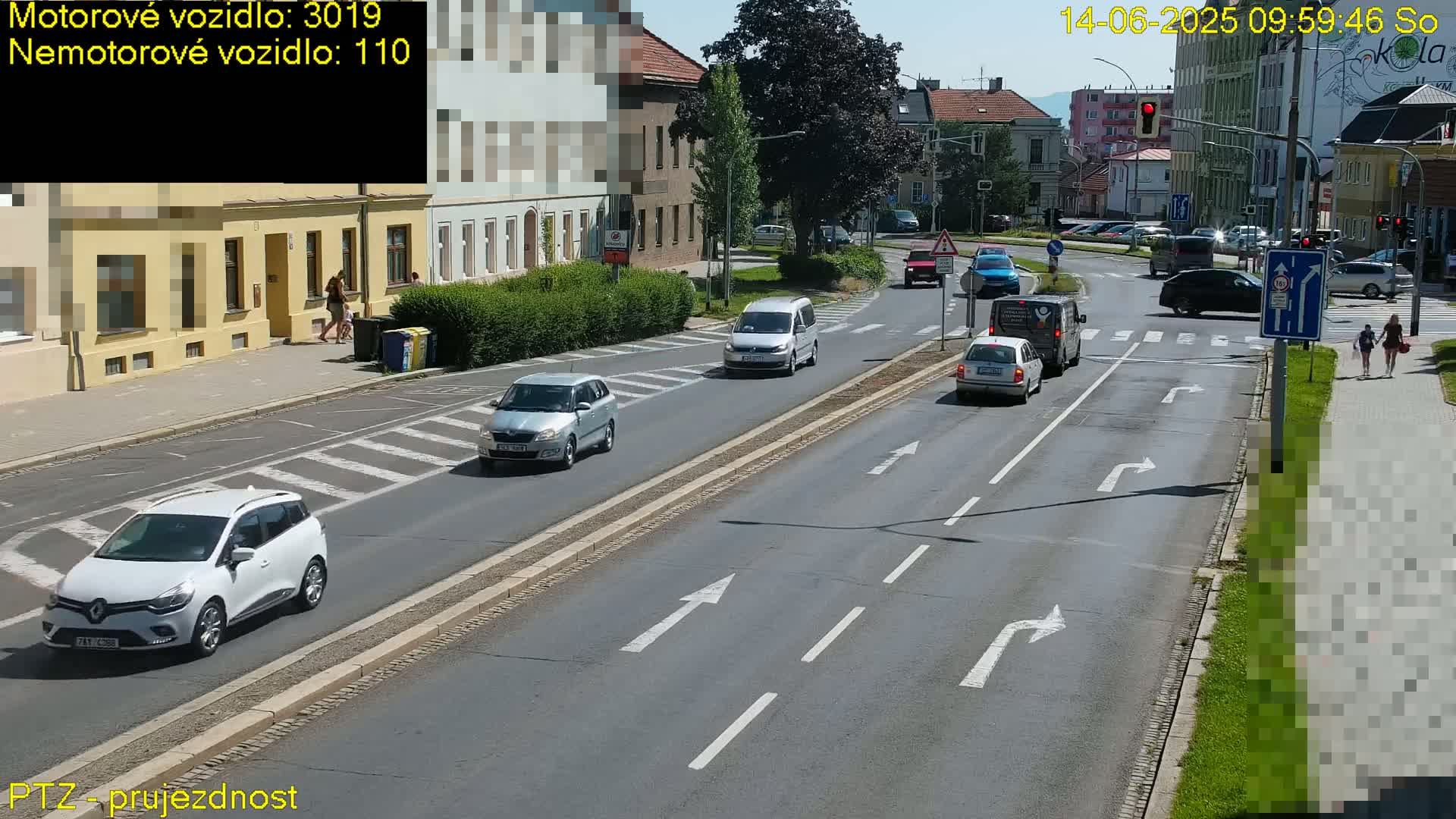 A sunny day shows several vehicles driving on a multi-lane road that intersects with another road, pedestrians walking on sidewalks, and buildings lining the street.