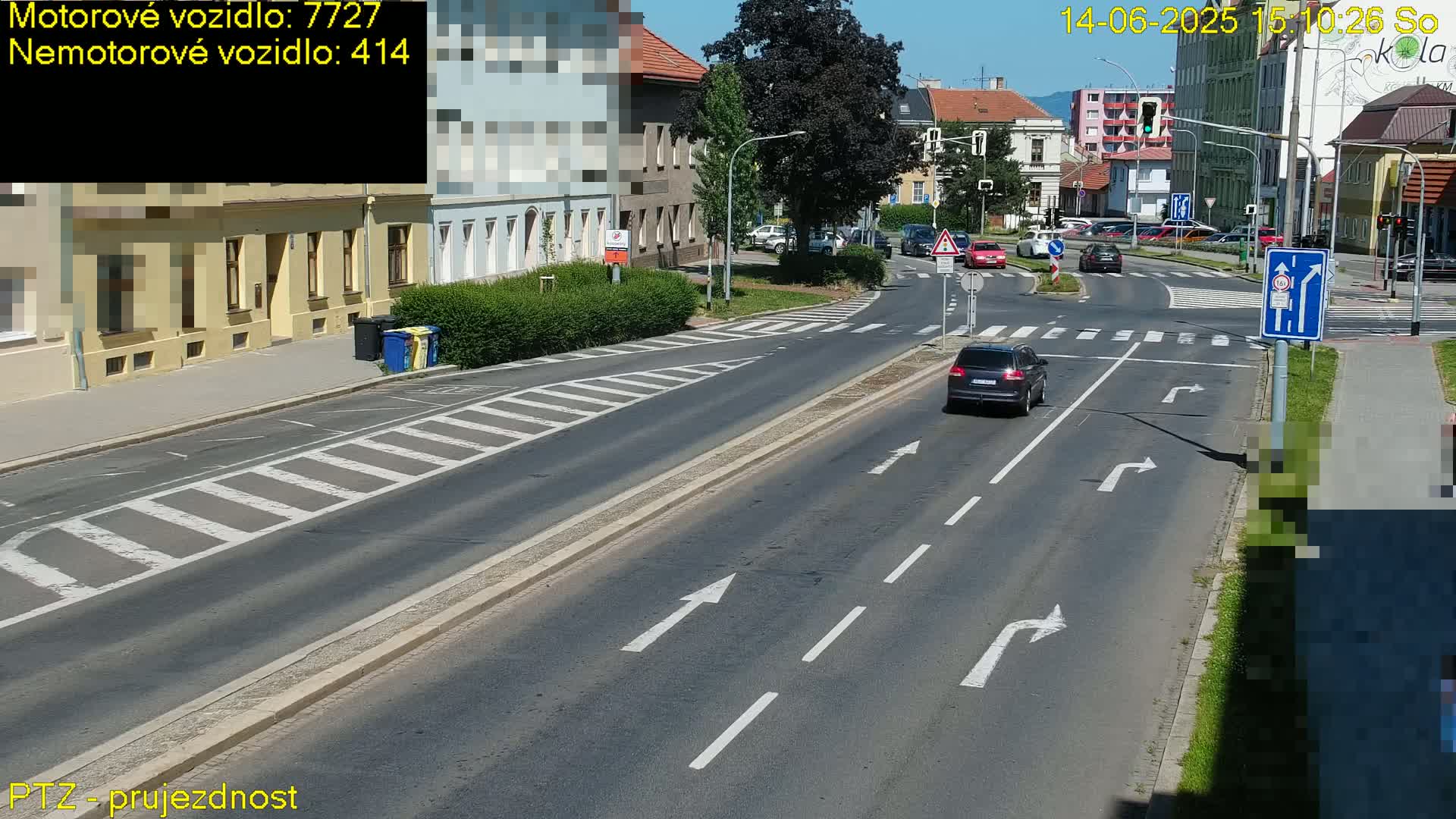 A sunny day shows a dark-colored SUV driving on a multi-lane road near a pedestrian crossing, with buildings and parked cars in the background.