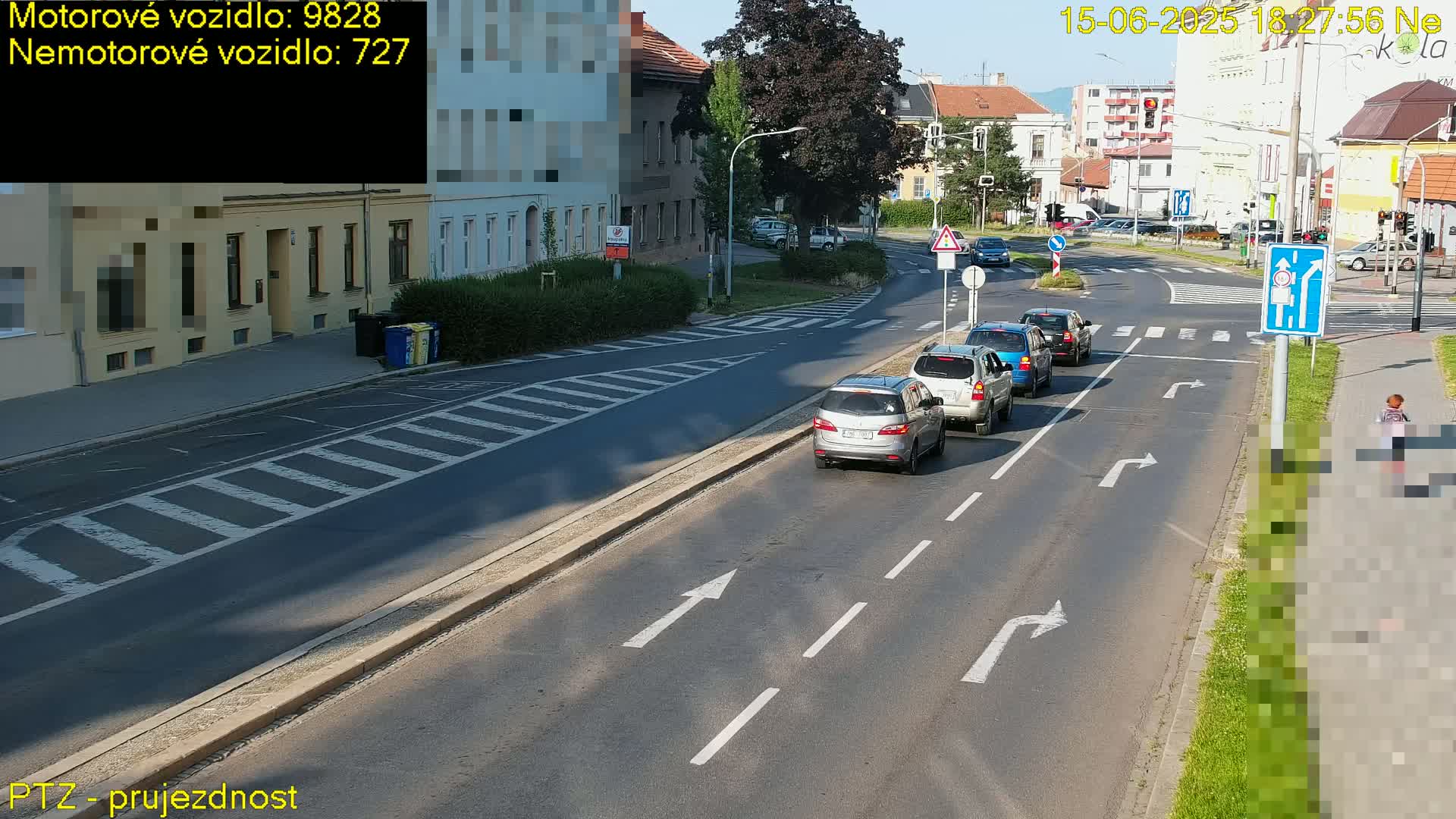 A sunny day shows several cars driving on a multi-lane road, with a pedestrian walking on the sidewalk next to a crosswalk.
