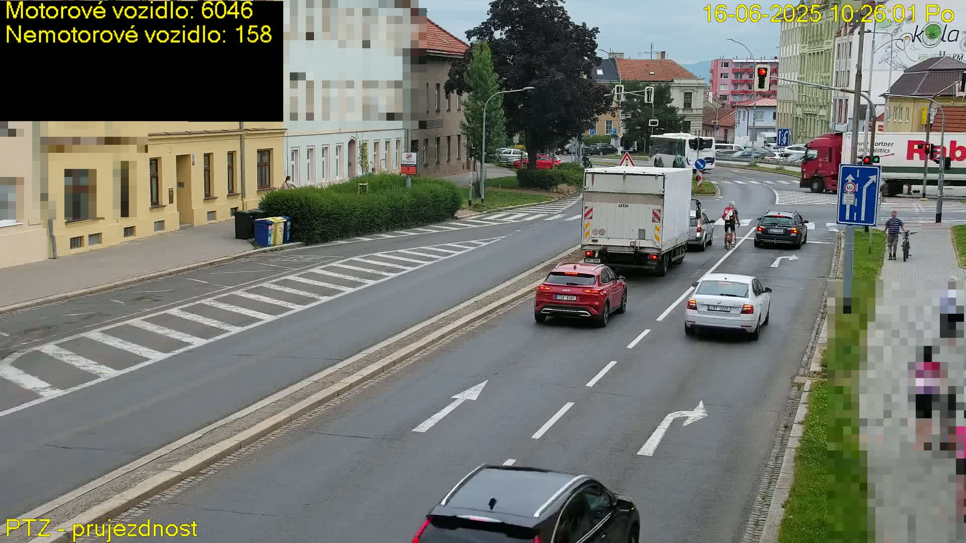 A street scene on a partly cloudy day shows several cars and trucks driving on a road with pedestrian crossings, alongside a sidewalk where people are walking and cycling.