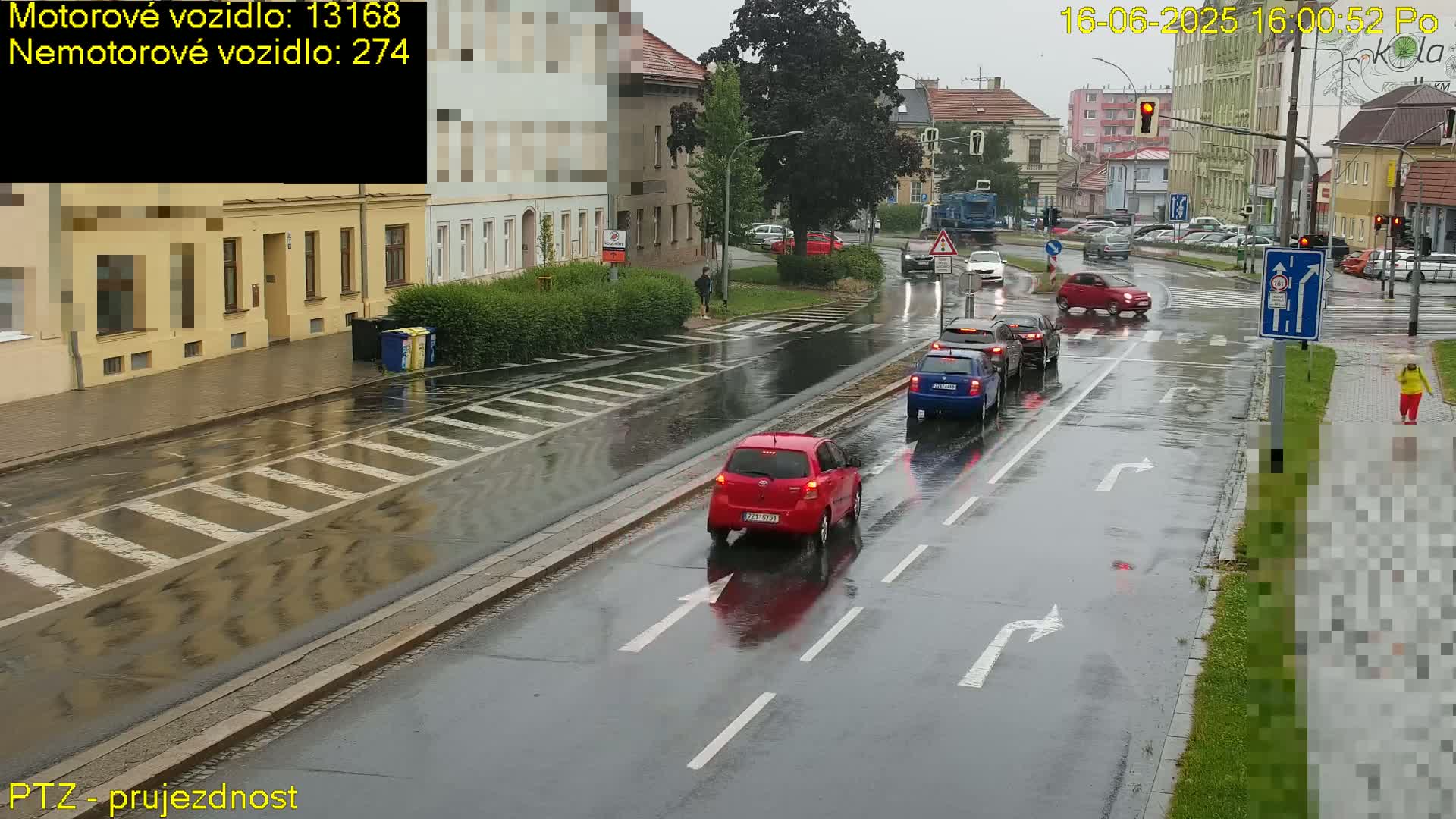 A rain-slicked city street shows several cars driving past a pedestrian crossing, with buildings lining the street on both sides.