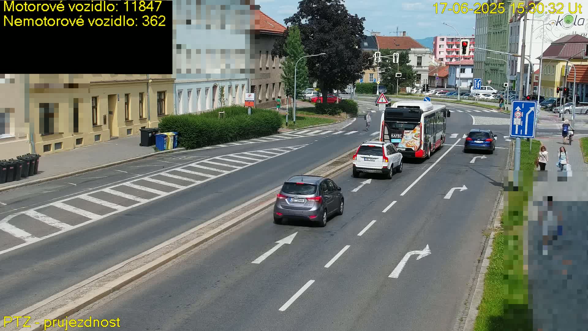 A sunny day shows several cars and a bus driving on a multi-lane road that intersects with a crosswalk, where pedestrians are walking.
