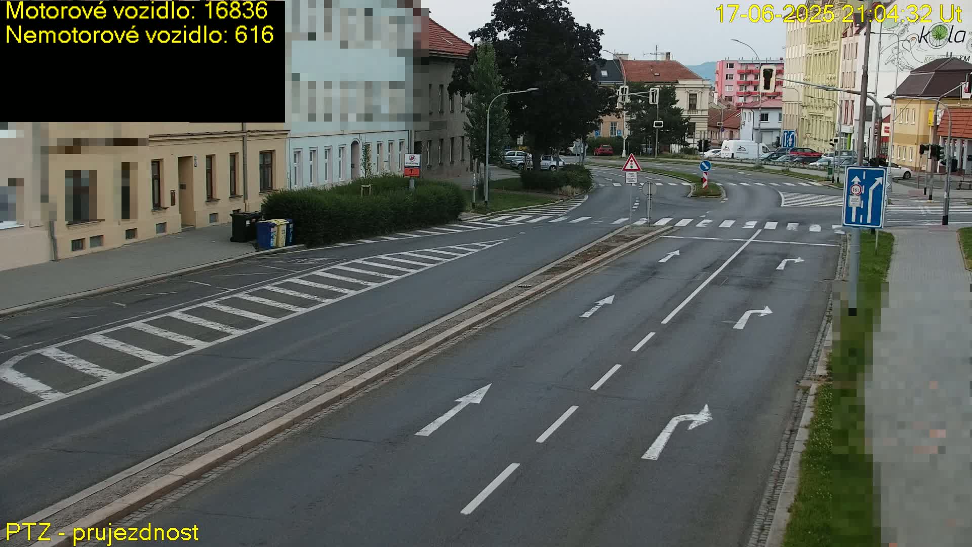 A wide, multi-lane road intersection with pedestrian crosswalks, bordered by buildings and greenery under a seemingly overcast sky.