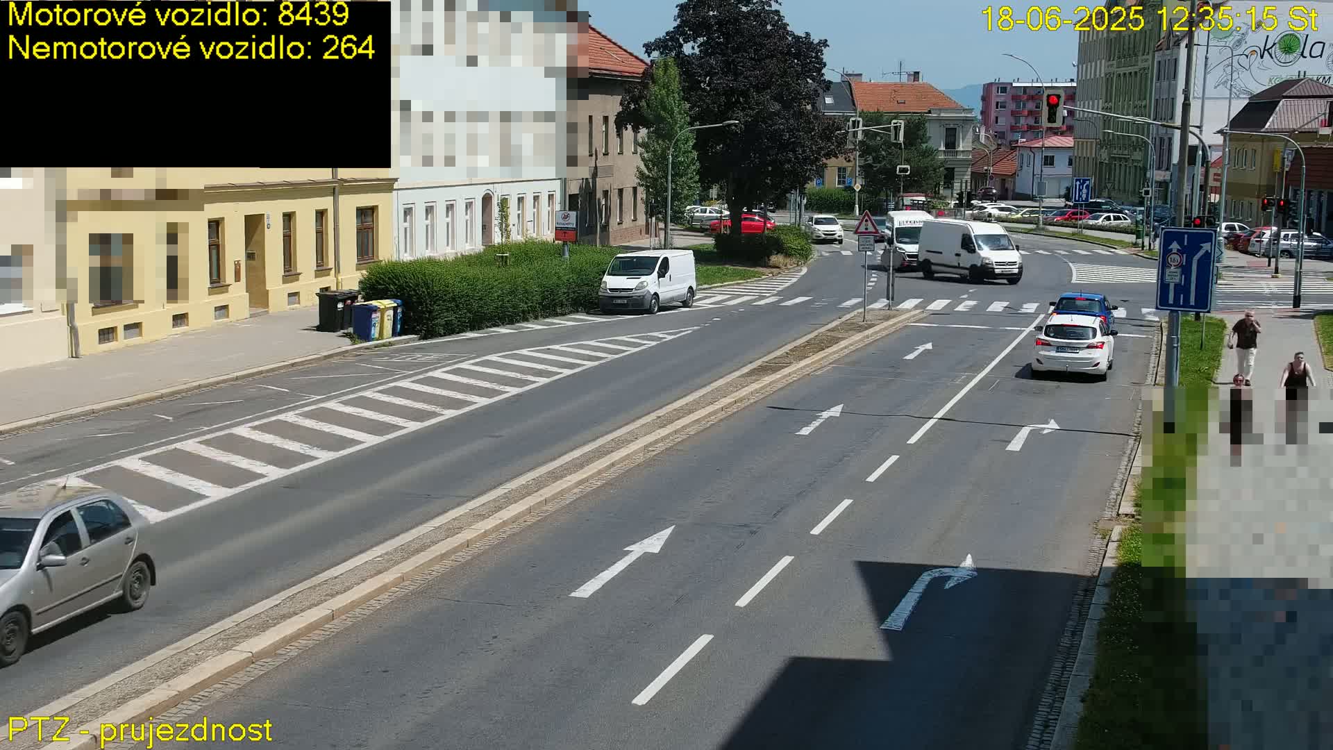 A sunny day shows a multi-lane road intersection with several cars, vans, and pedestrians, some crossing a crosswalk.
