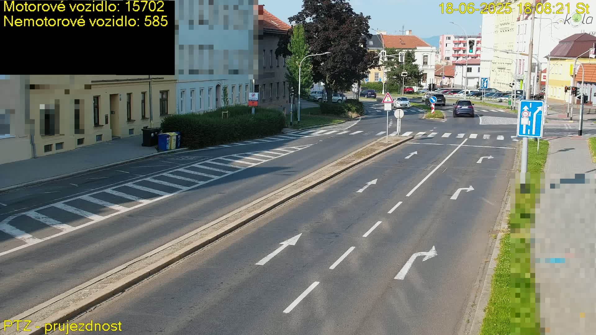 A sunny street scene shows a multi-lane road with marked lanes and crosswalks, several cars, and buildings lining the street.