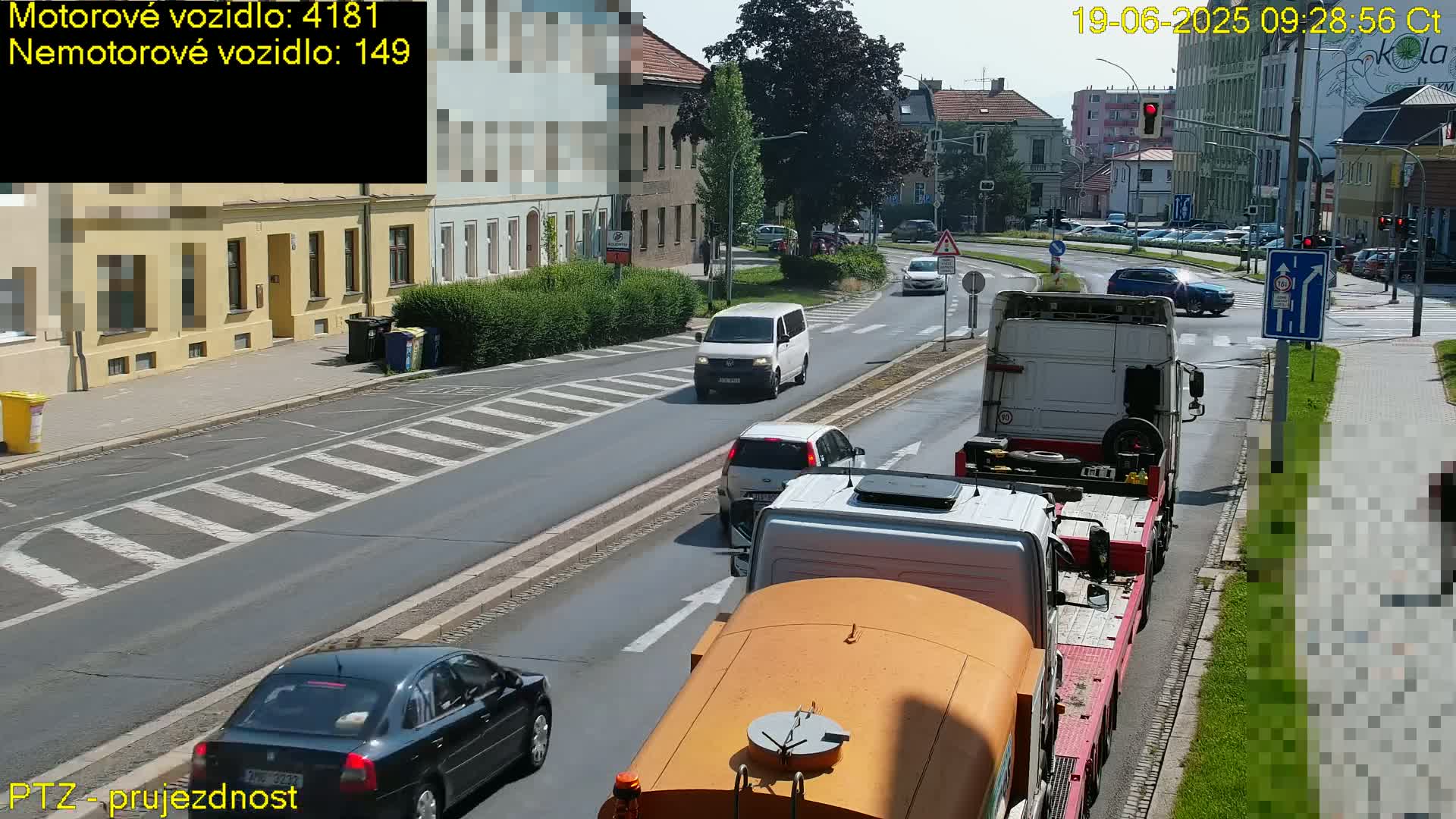 A sunny day view of a multi-lane road with several cars and trucks, including a large flatbed truck hauling another truck,  near a pedestrian crossing.