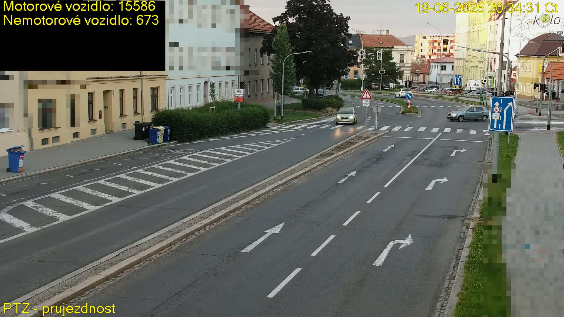 A street scene at dusk shows a multi-lane road with several cars and a crosswalk, with buildings and hedges lining the street.
