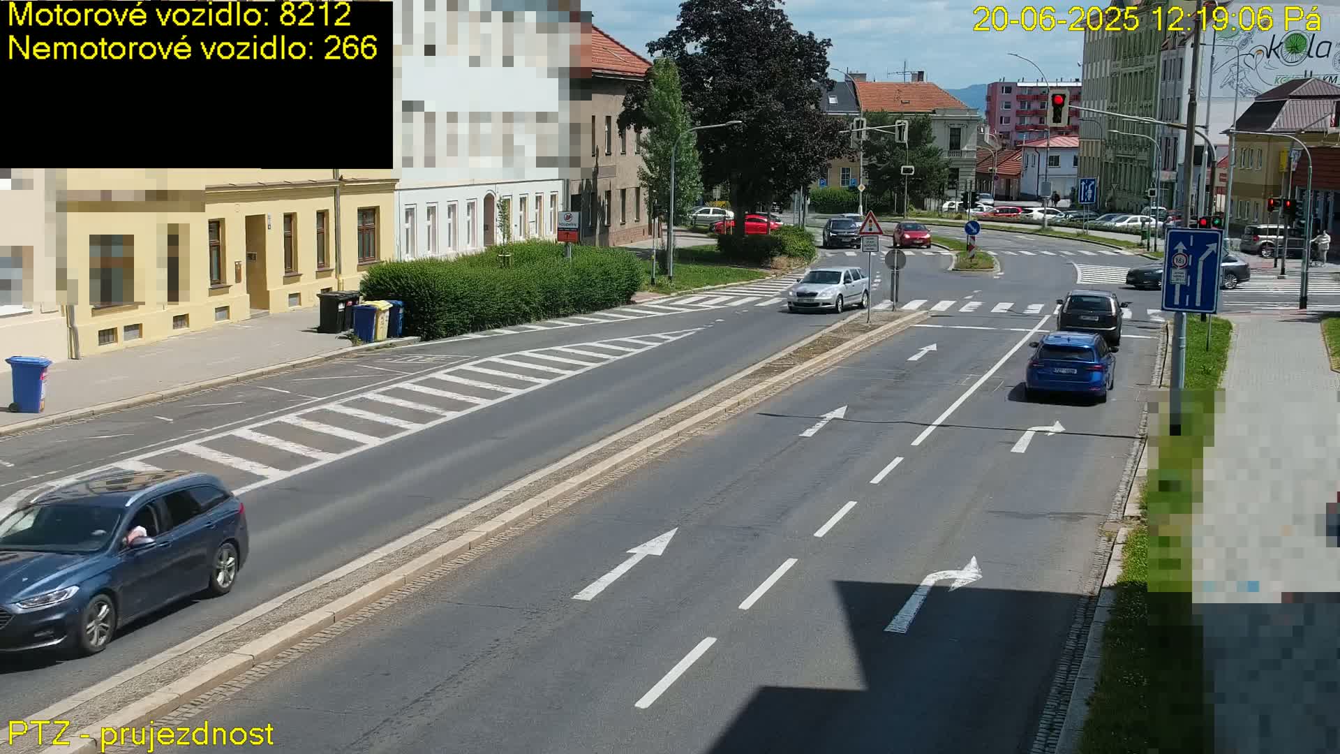 A sunny day shows several cars driving on a multi-lane road with crosswalks and buildings lining the street.