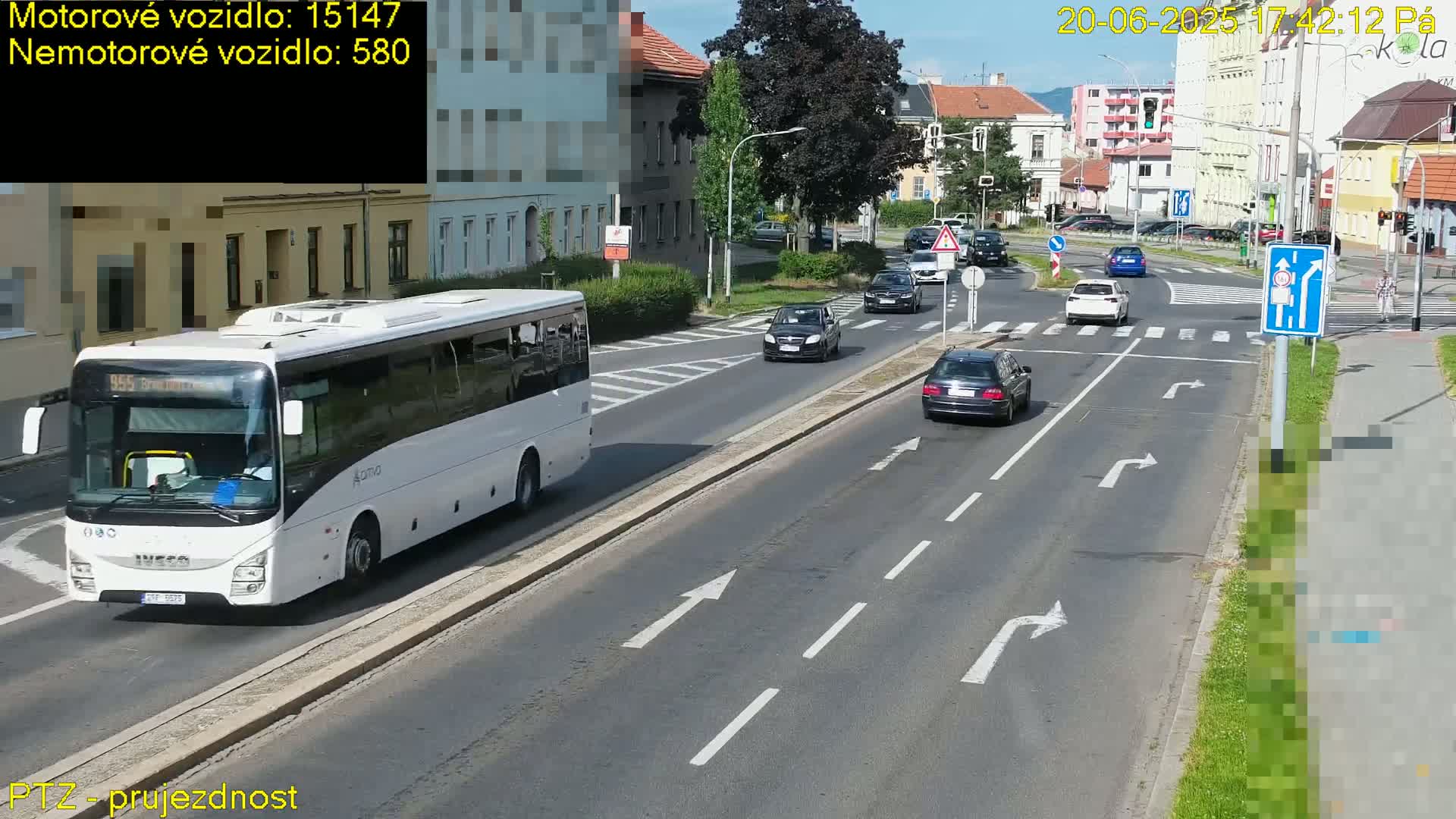 A white bus and several cars are driving on a road on a sunny day, passing through a crosswalk at an intersection near buildings.