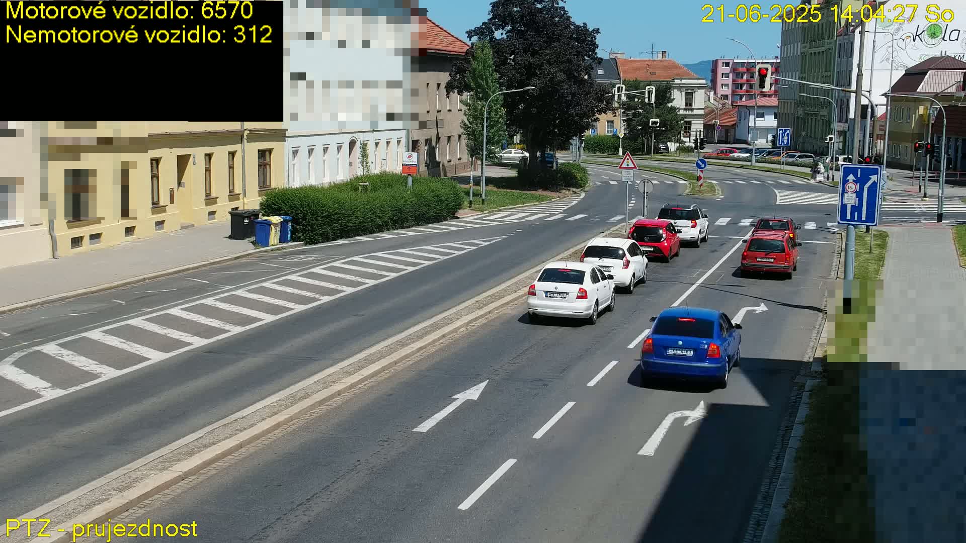 Several cars are driving on a sunny city street with crosswalks and buildings lining the road.